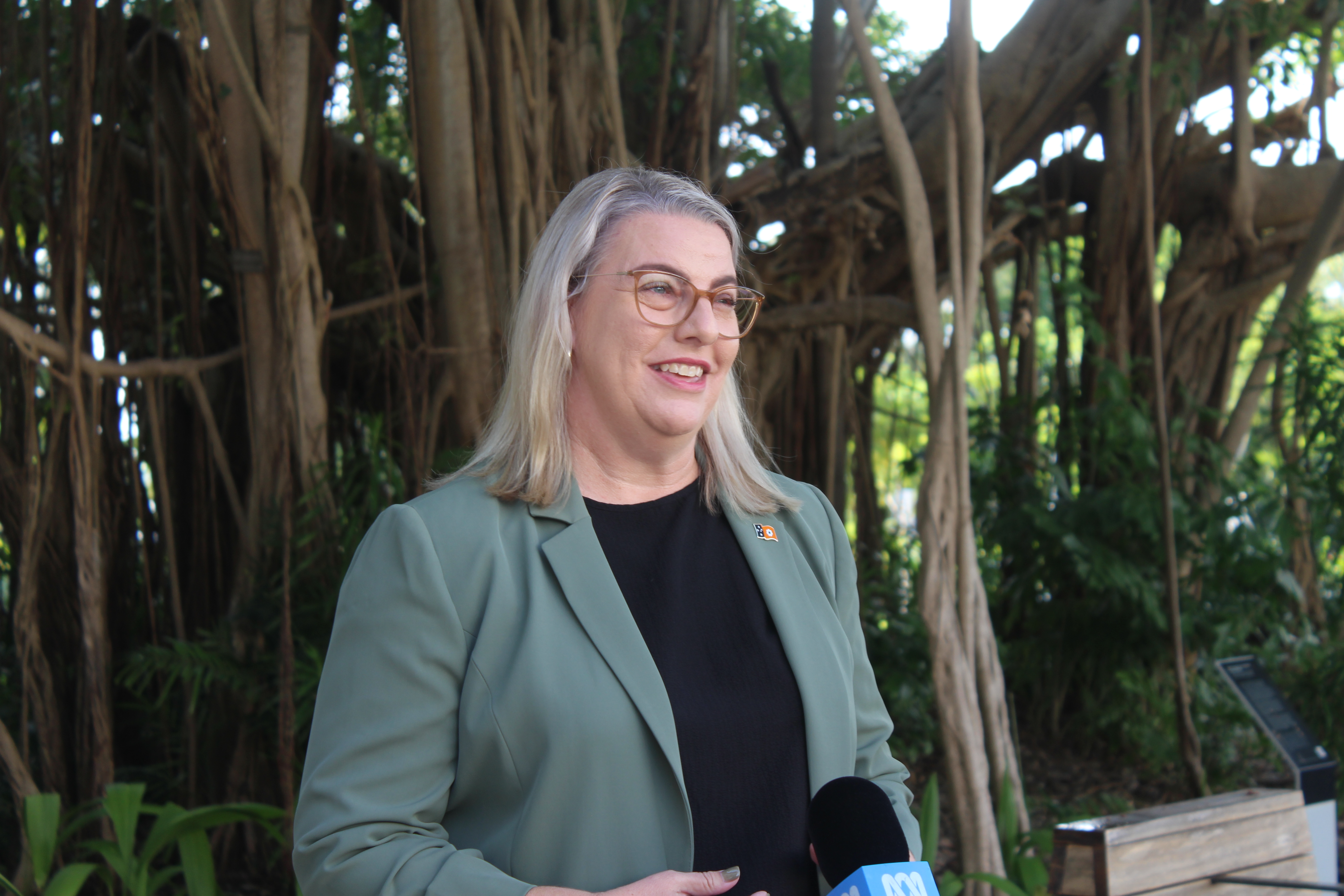 Blonde woman in sage-coloured jacket stands outdoors in front of a press conference microphone. 