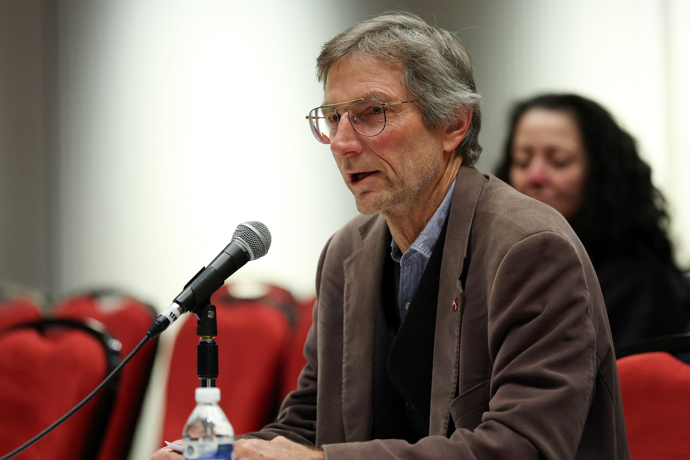 Man with glasses and brown jacket speaks into microphone.