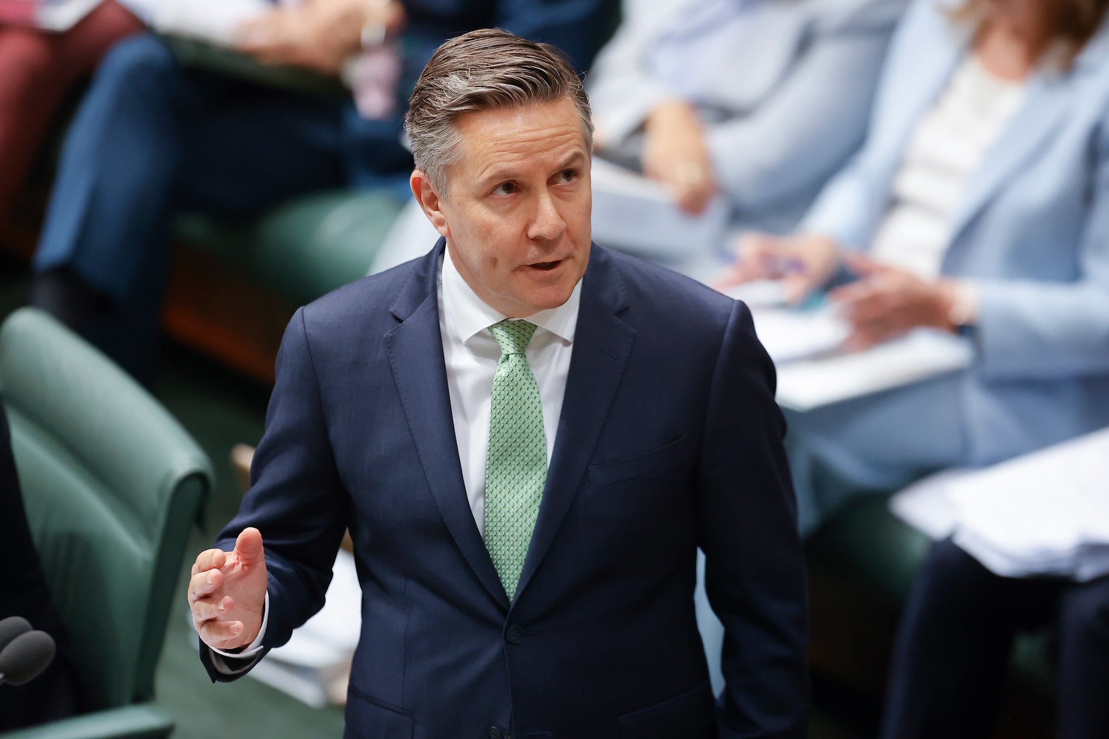 A man in a suit with a green tie speaks in the Australian parliament. Seated individuals in business attire are blurred in the background.