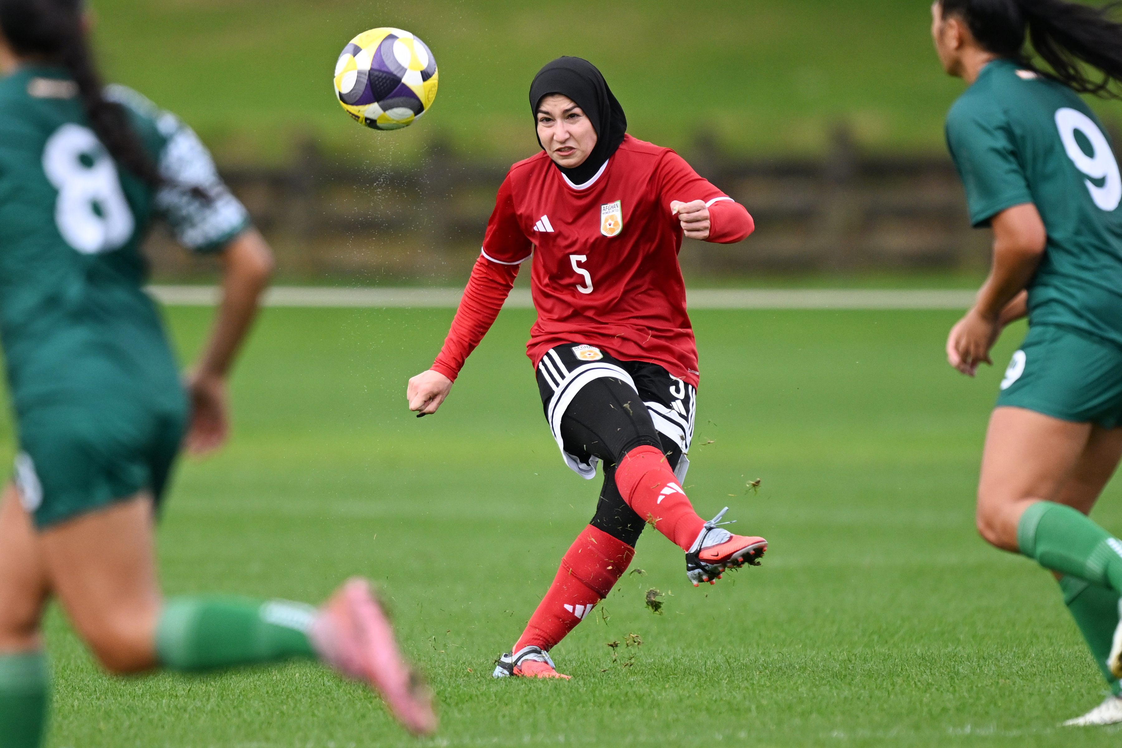 Young woman in a chador and soccer uniform kicks a ball in a game between Afghan Women United and Cook Islands.
