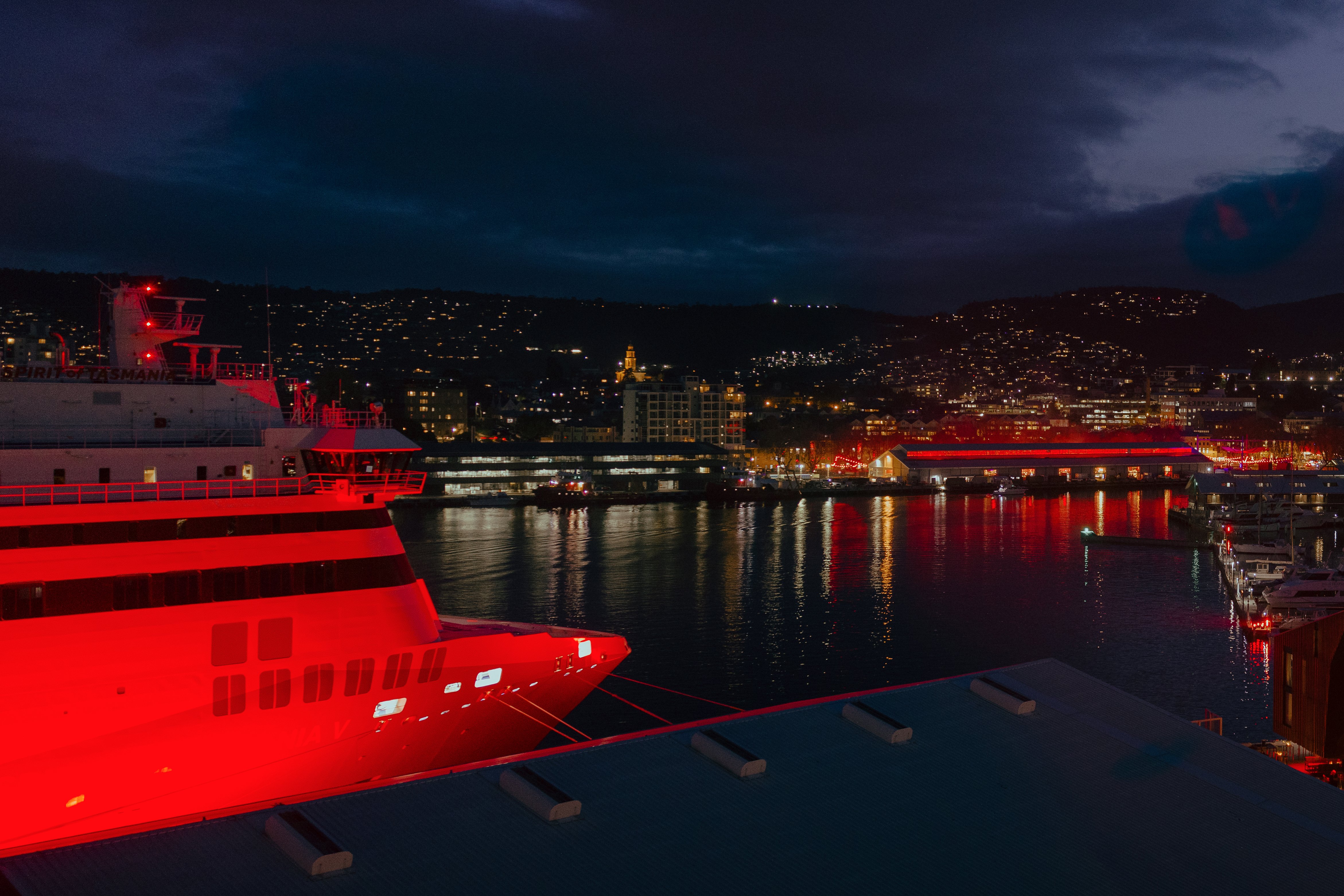A large cruise ship illuminated in red is docked in a harbour at night. City lights reflect on the water, with dark, clouded skies above.