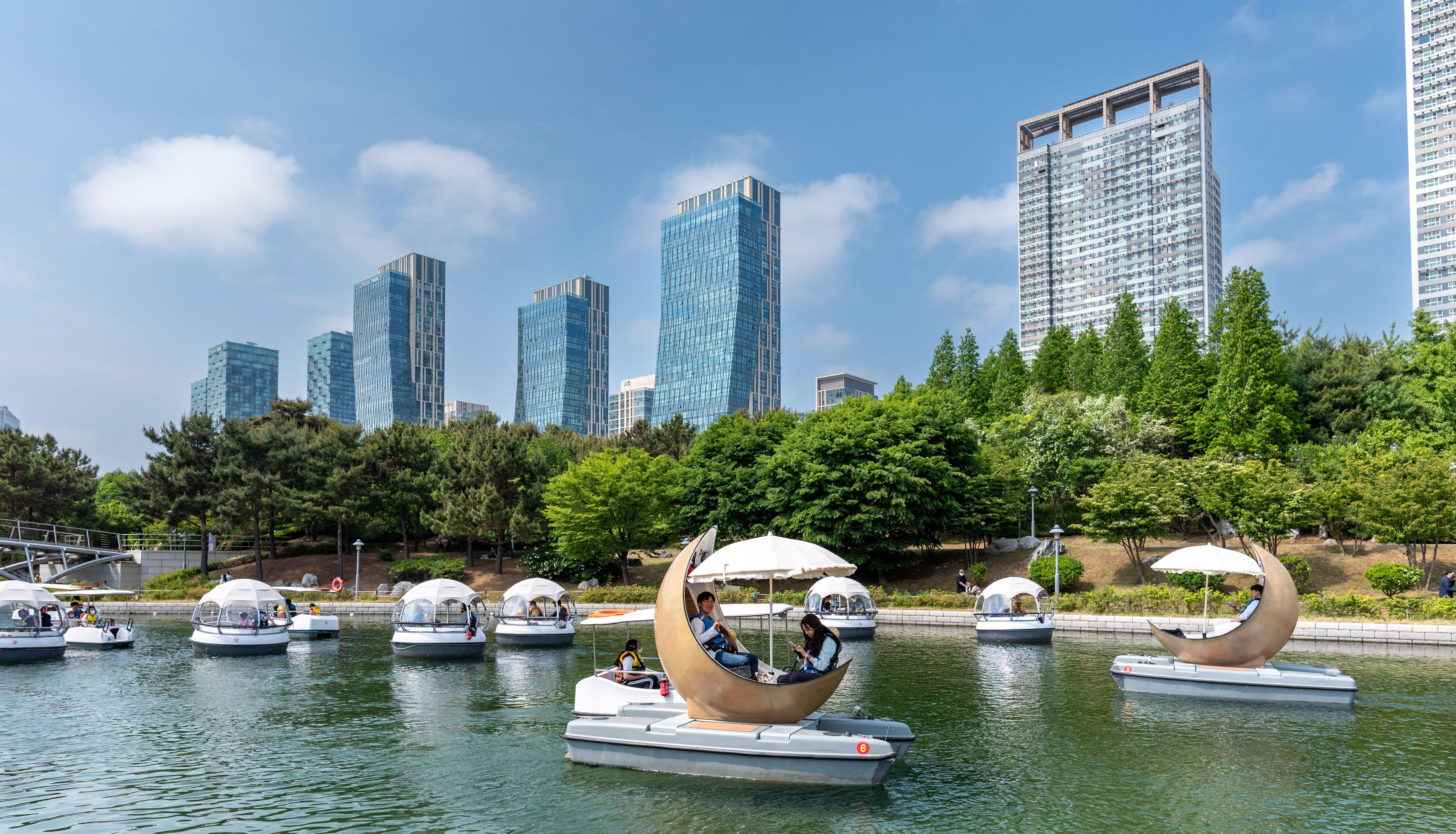 People floating on boats in a park lake with city skyscrapers in the background.