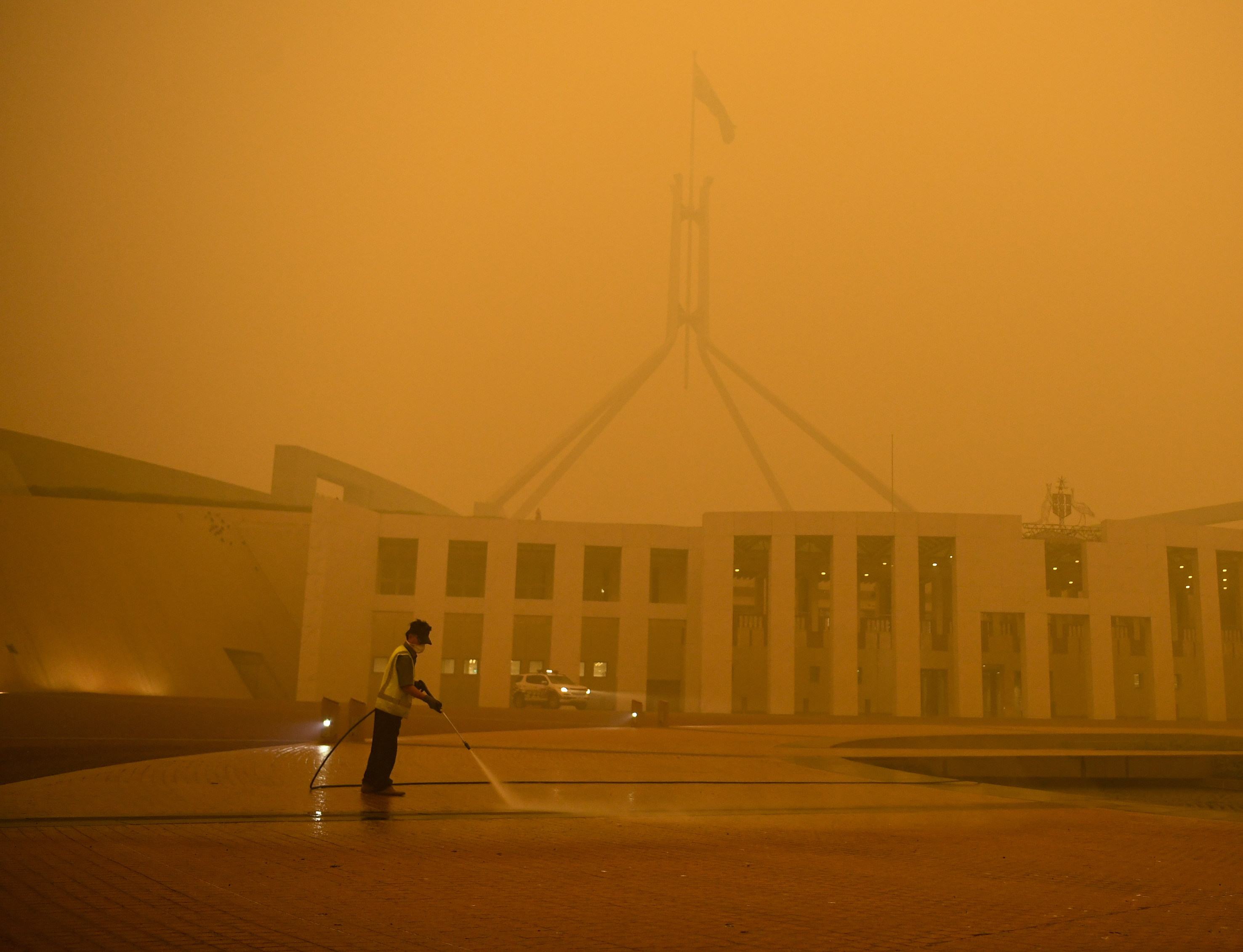 A cleaner wearing a face mask stands in the forecourt of Parliament House, Canberra They are surrounded by an orange haze caused by bushfires.