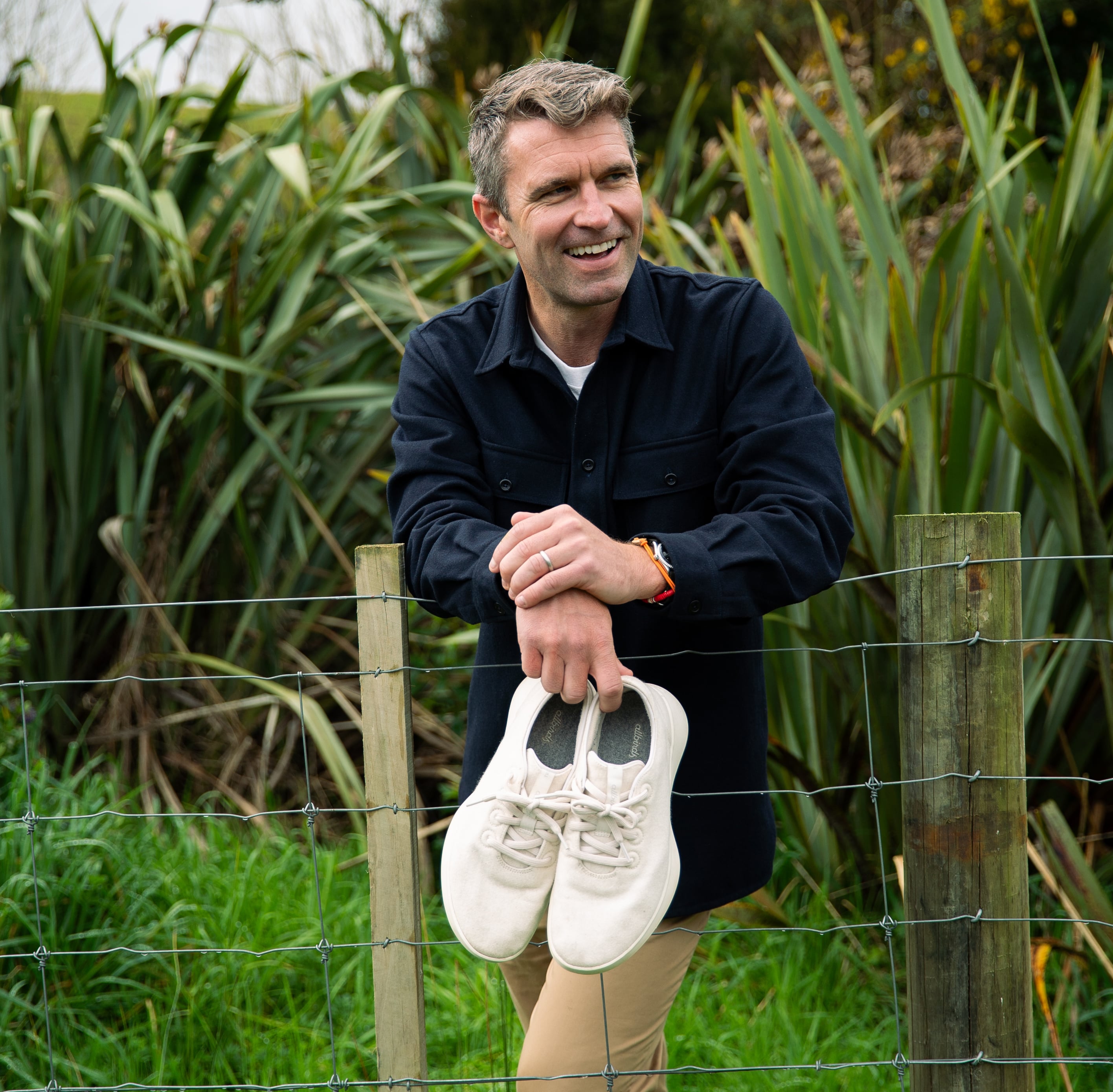 A man holding a white pair of shoes as he leans on a wire fence.