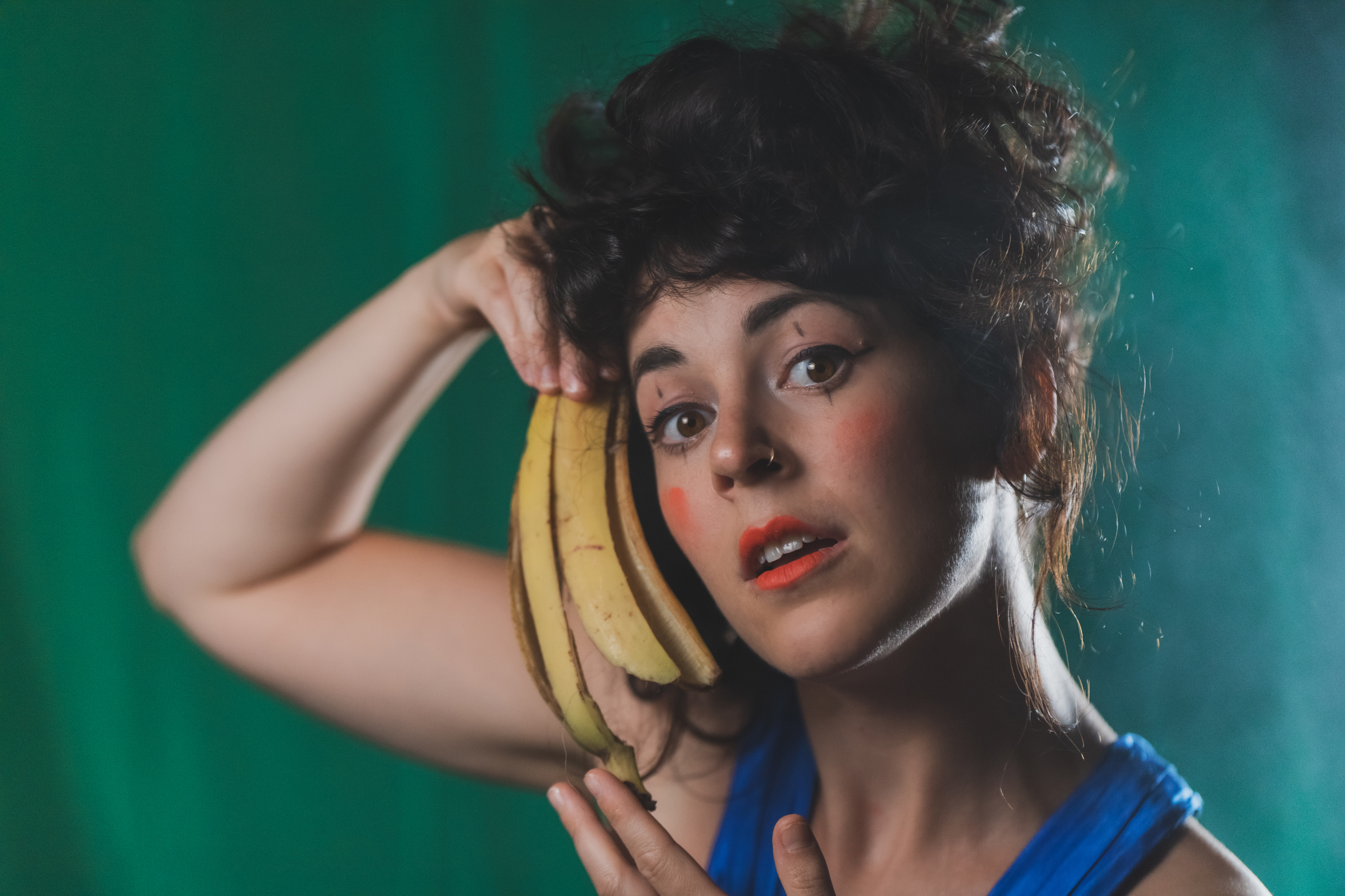 Performer Britt Plummer is in make-up and holds an empty banana peel to the side of her face as she gazes into the camera.