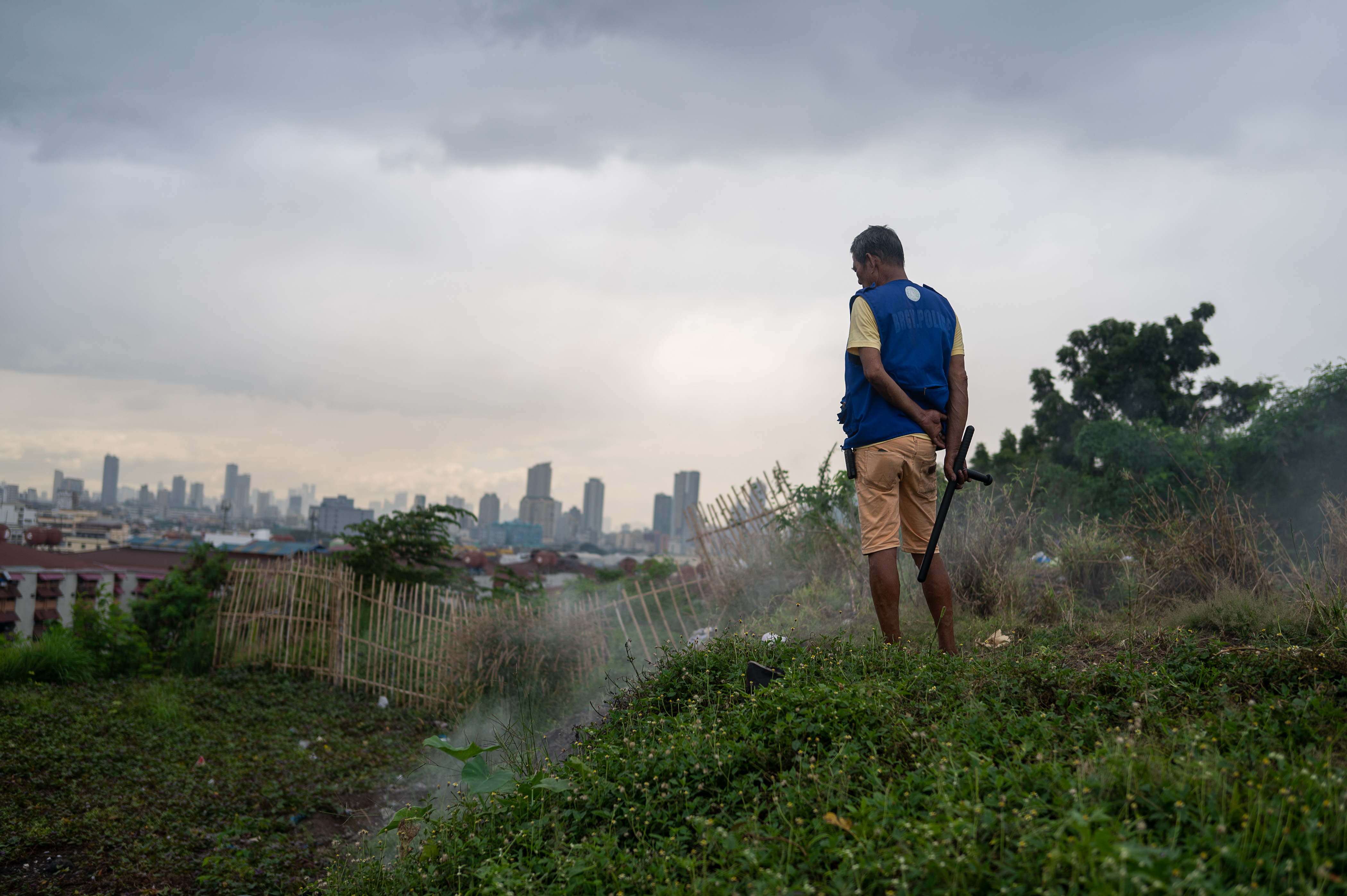 A man wearing shorts and a vest, carrying what looks like a baton stands on a green hill filled with flowers. Beyond him is a rickety fence and a city skyline.