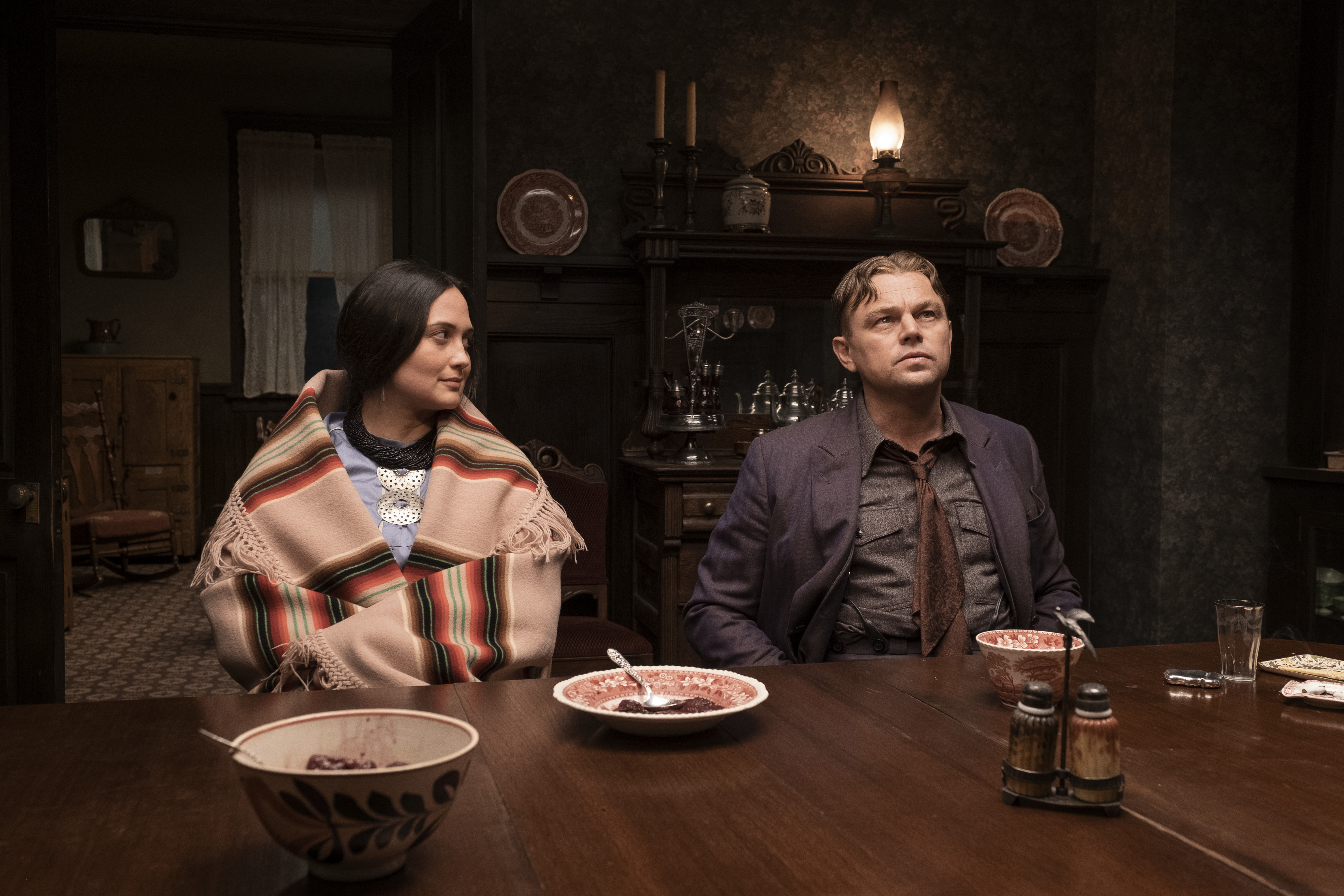 A woman wrapped in a blanket sits with a man wearing a blue blazer at a dinner table.