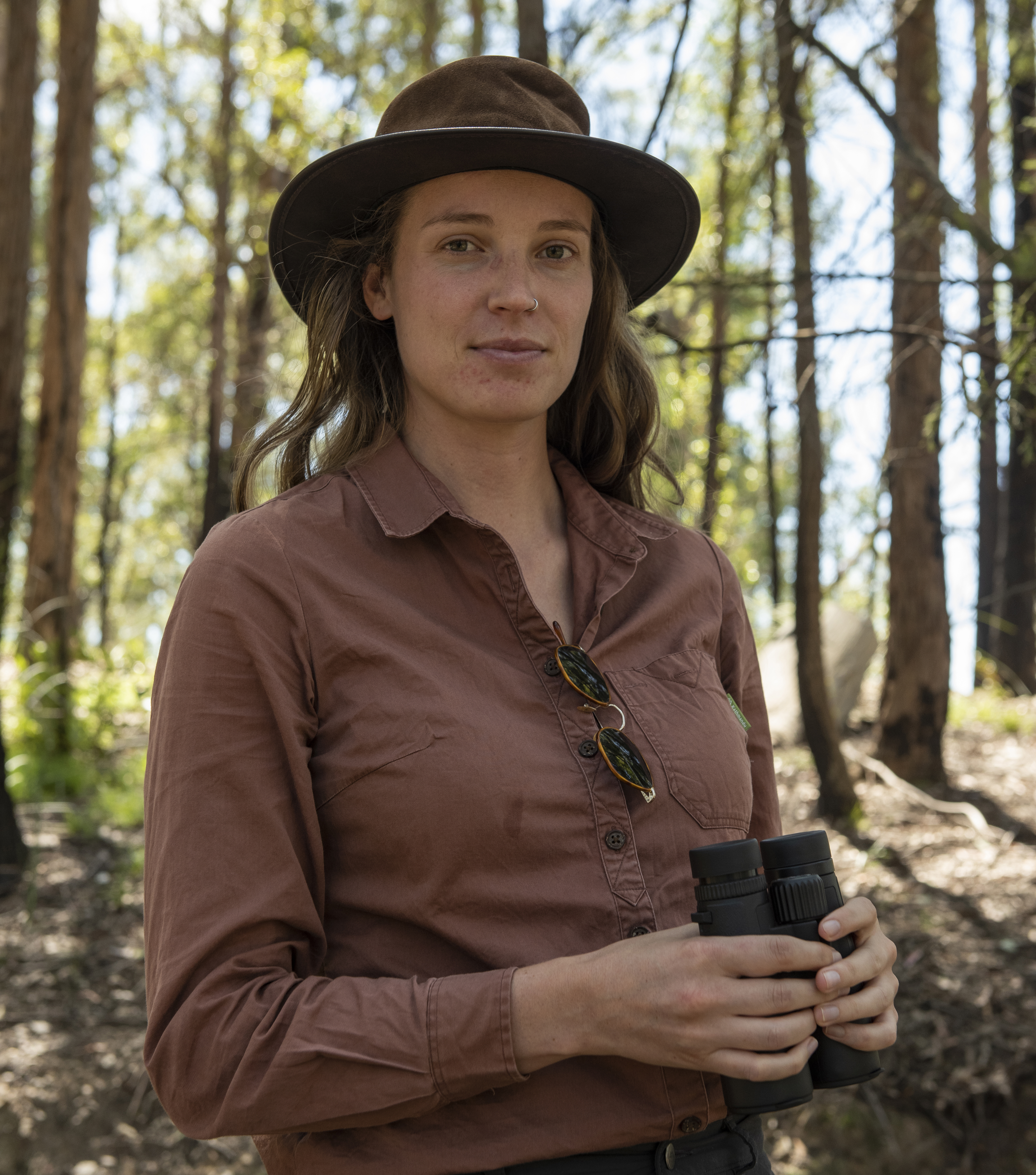 Portrait image of a woman in a brown shirt and wide-brim hat standing in a forest.