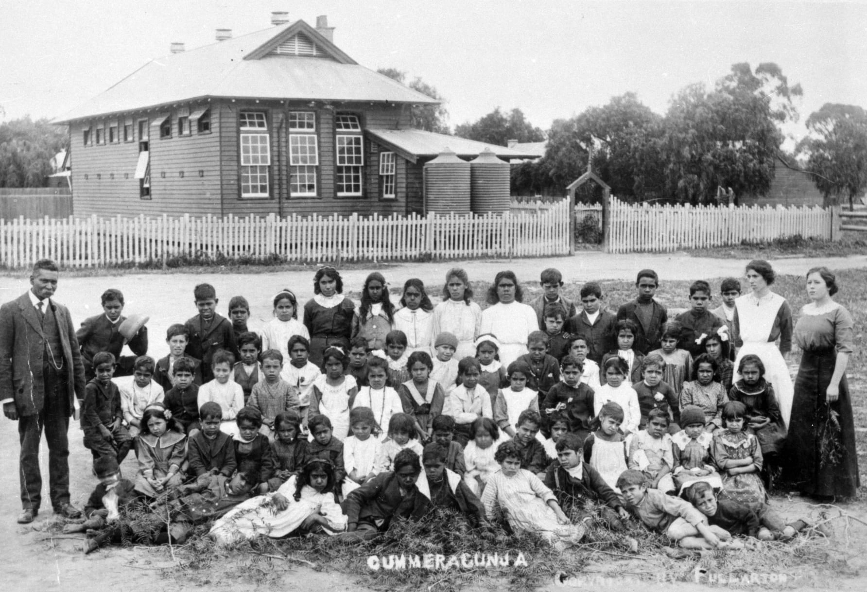 A black and white photograph of a class of school children with their teacher. The school is in the background.
