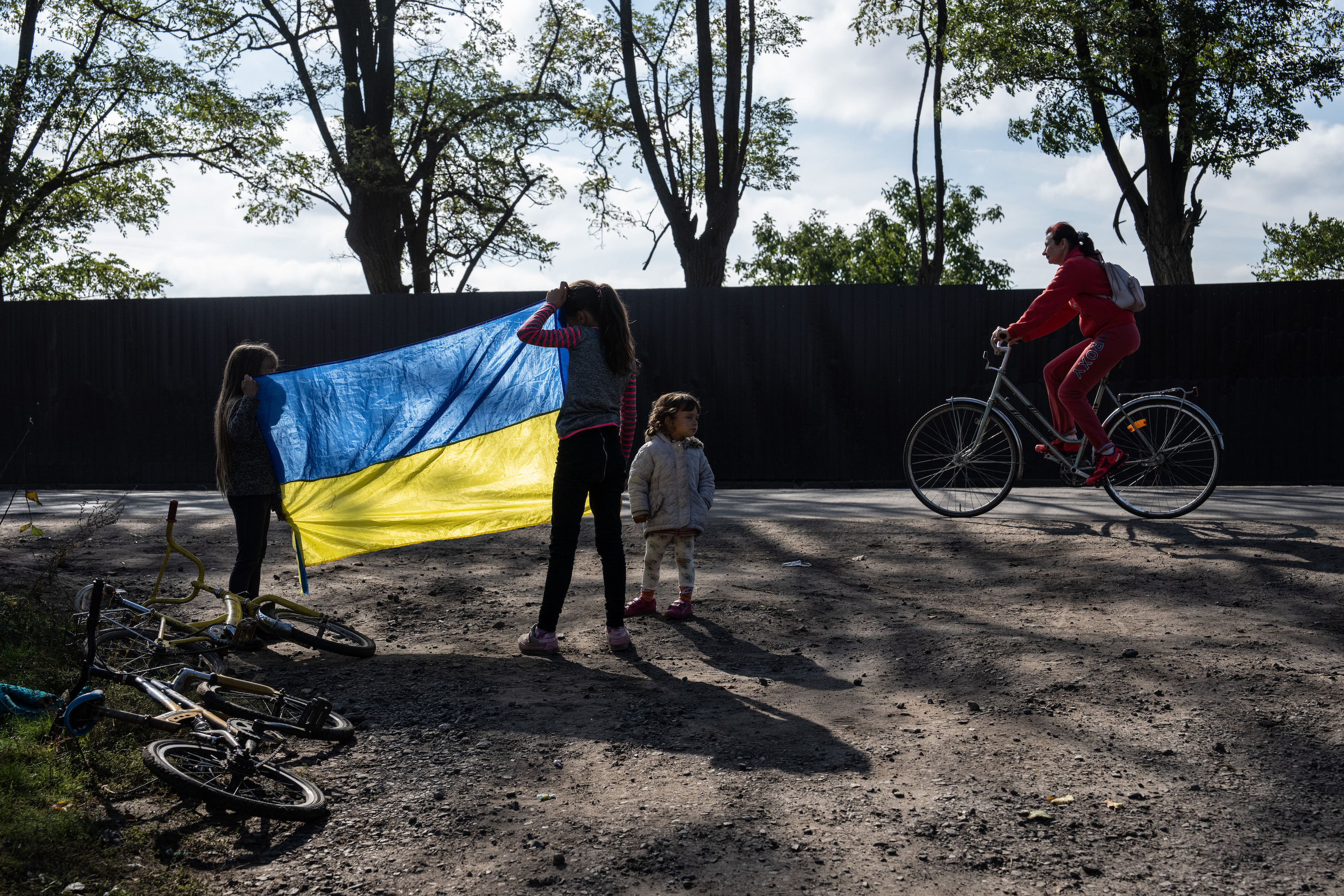 Two young girls hold up a blue and yellow Ukrainian flag on a gravel road. There are two bikes on the ground near them, a smaller child next to them and a woman on a bike about to pass by. The image feels sad and heavy.