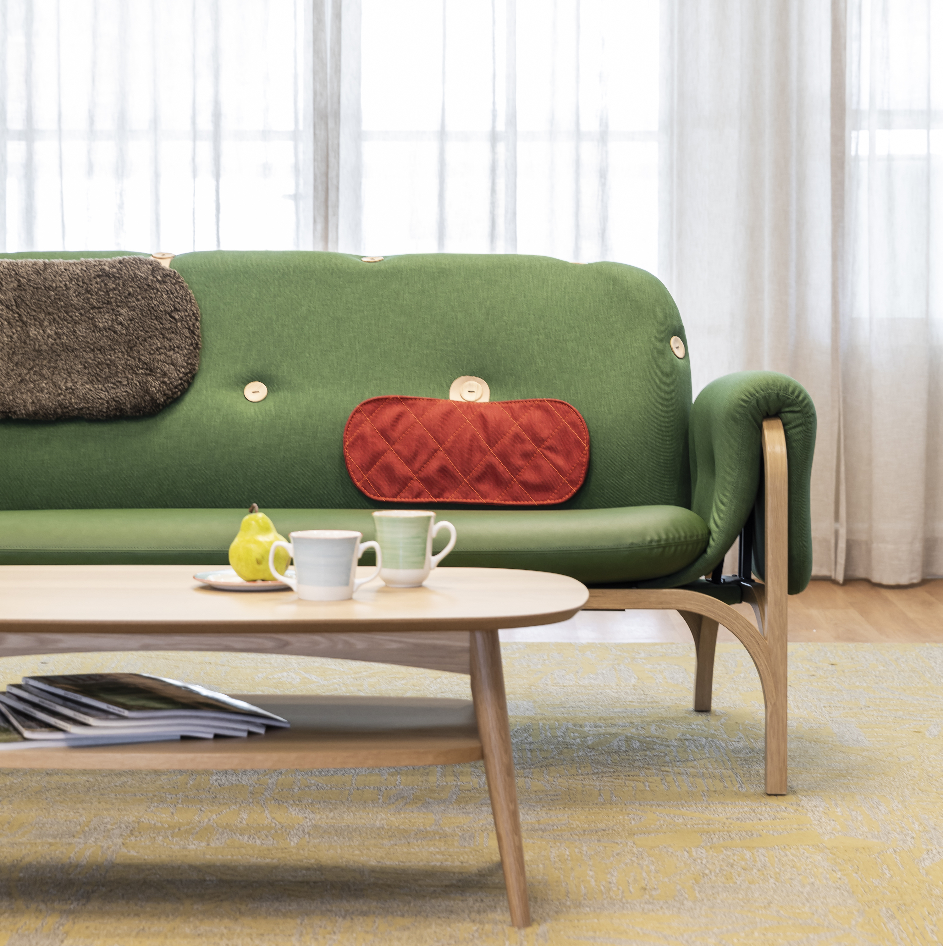 A clean modern living room is shown, with a green upholstered wooden couch with red and grey cushions, and a simple wooden coffee table with two mugs and a pear, with a stack of magazines on the lower shelf