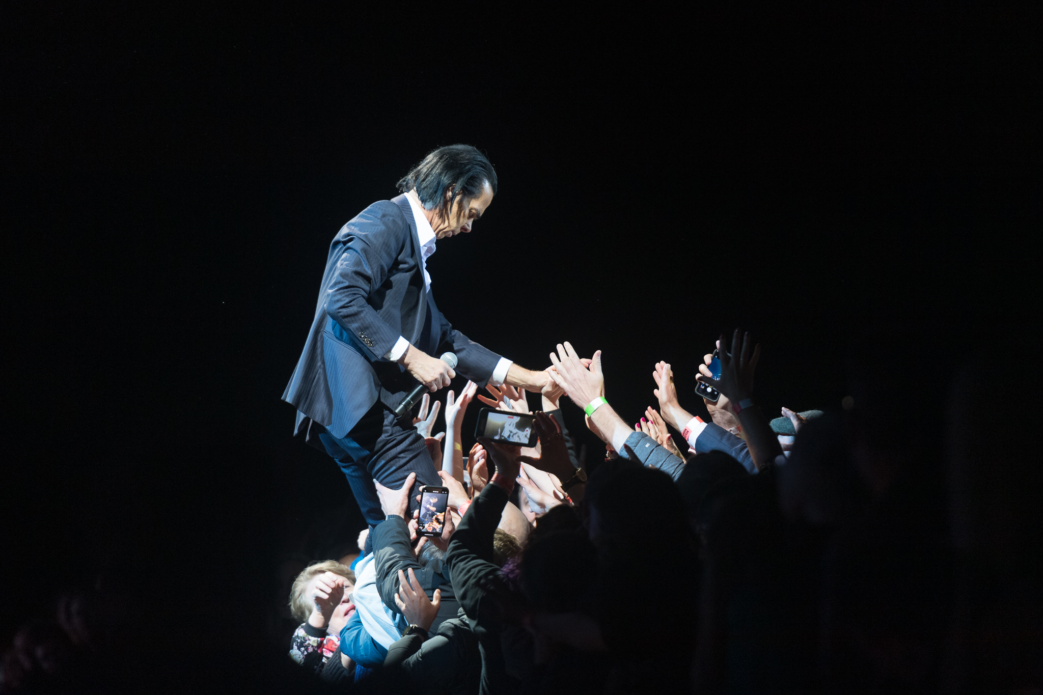 A white man in a dark suit with slicked back hair is on the very edge of a stage holding a microphone; he appears to be walking into the crowd in front of him as people reach out to hold him, some with mobile phones filming his movements