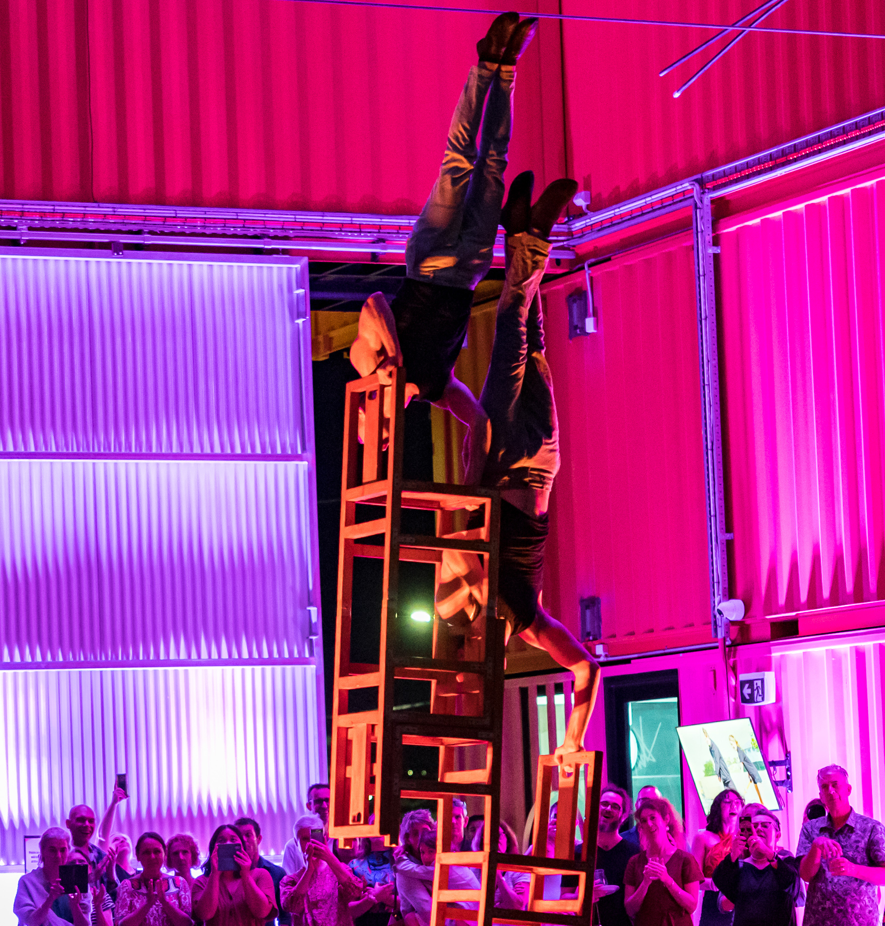 Two performers upside down on an abstract structure while an audience records on their phones.