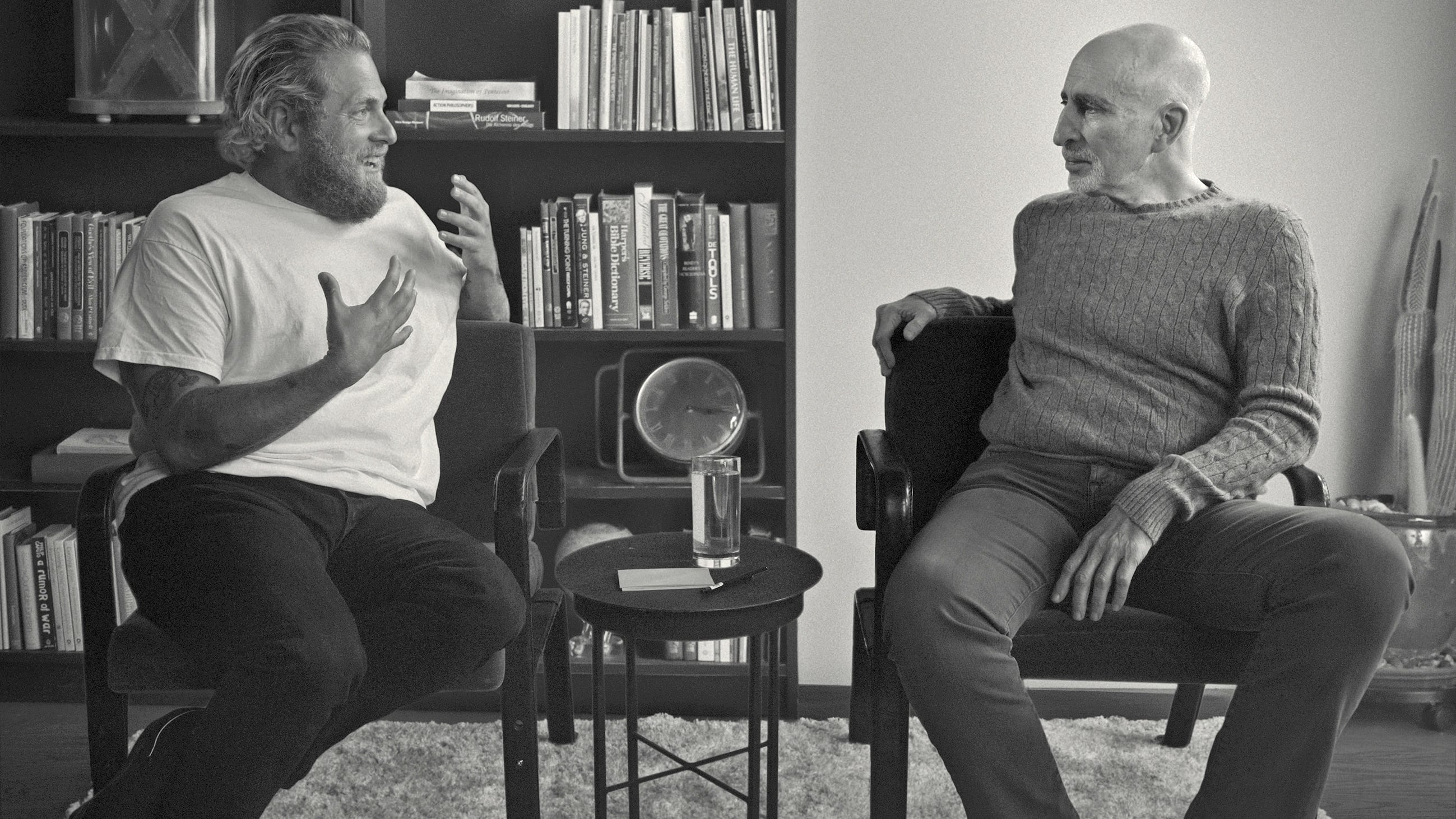 Two men are sitting on chairs next to each other in a simple library. One is younger, with a beard and wearing a white t-shirt, as he gestures while speaking. The other is older, gaunt and listens to the first speak. The image is in black and white.