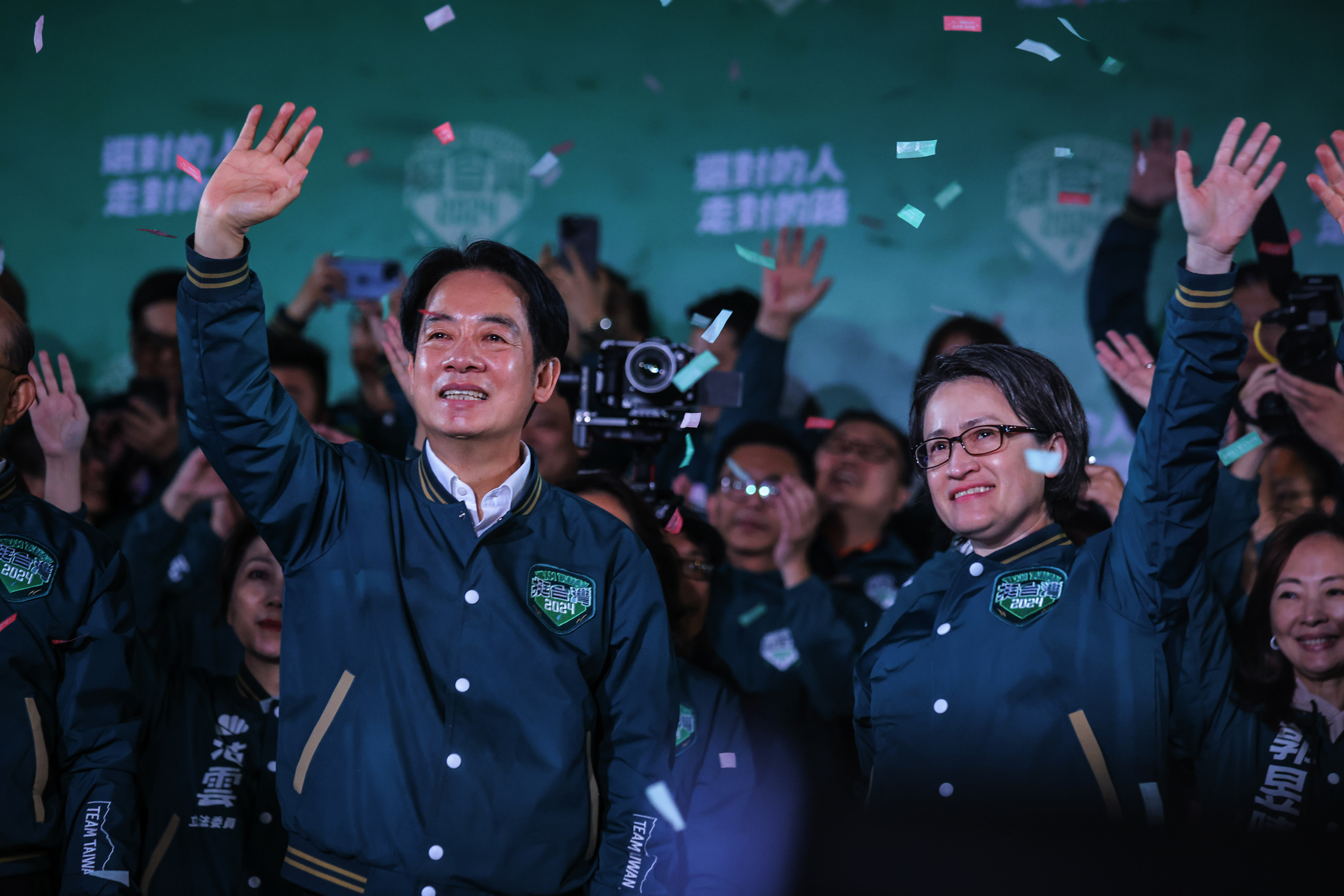 Politicians in bomber jackets wave to supporters as confetti falls around them.