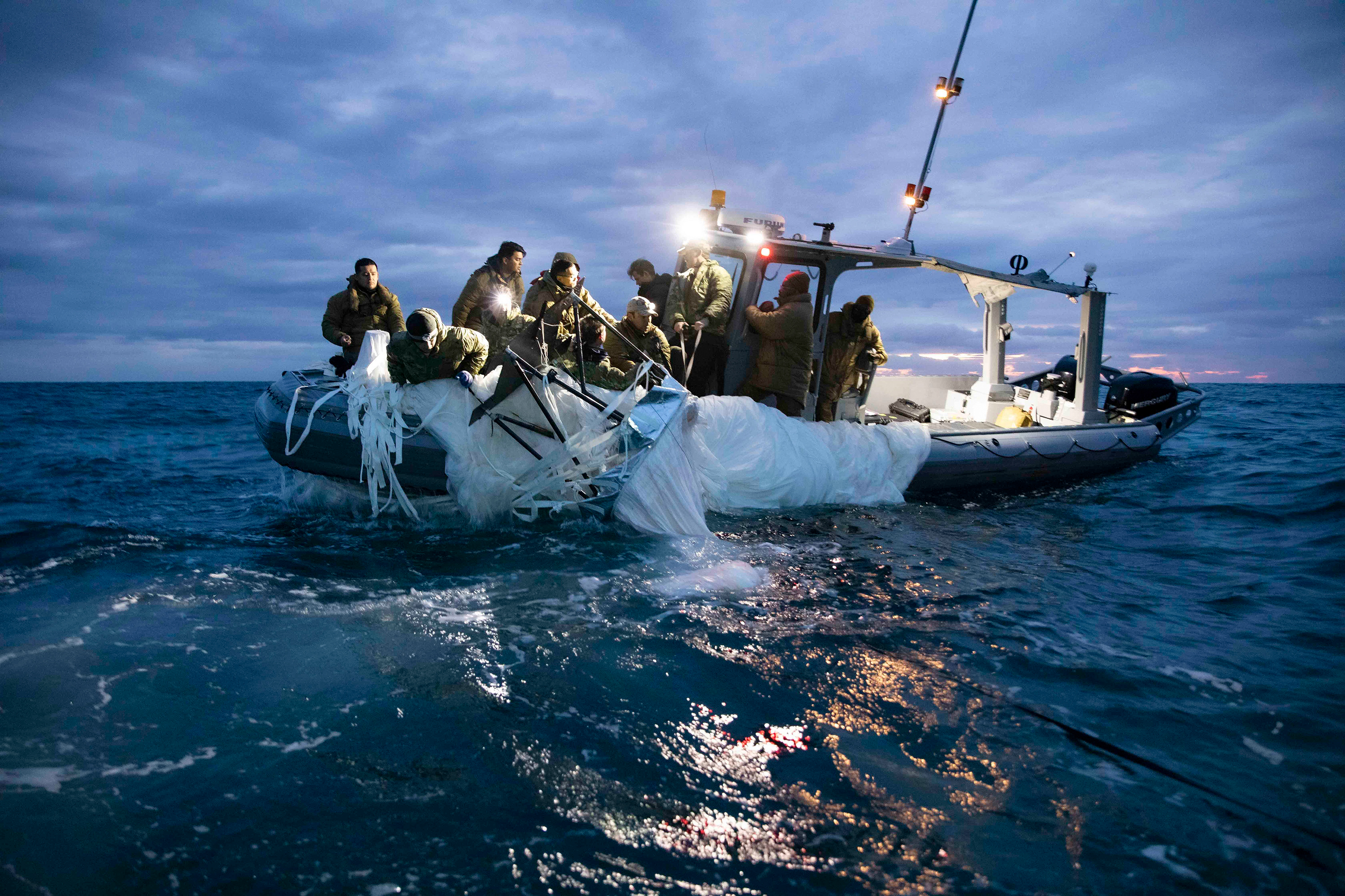 Sailors in thick jackets pull the rags of a balloon from the dark water. Their boat is illuminated by lights in the twilight.