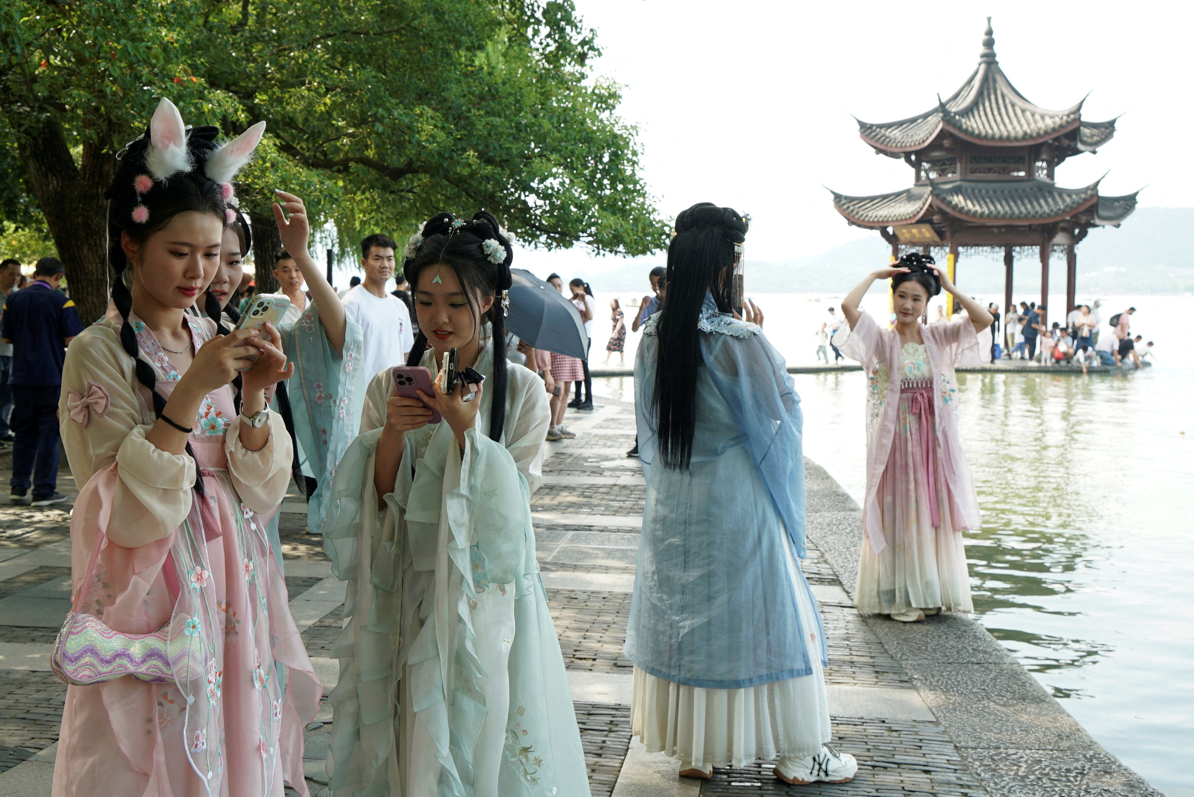 Women in traditional Chinese dresses on their phones as they stand beside a body of water.