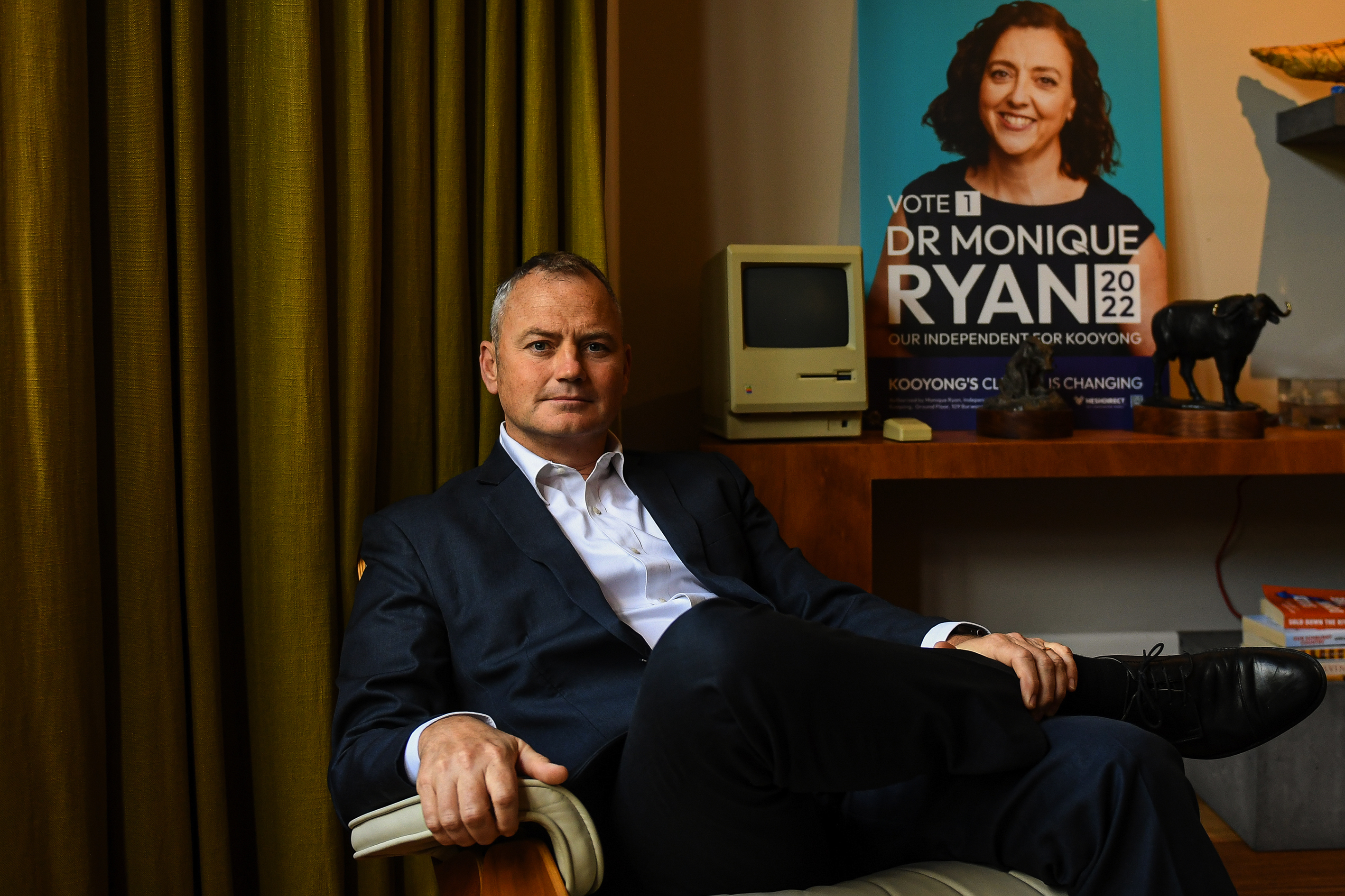 A man in a suit sitting in a chair beside a Monique Ryan poster.
