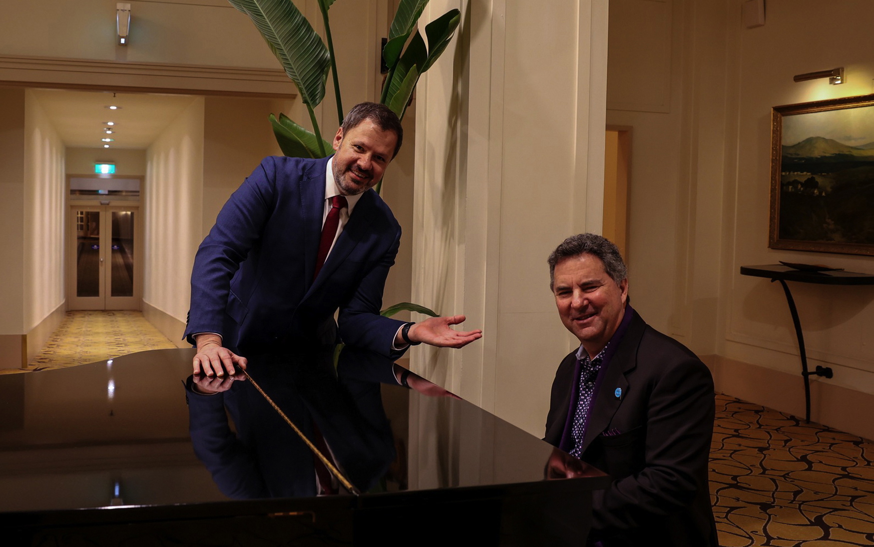 A man in a back blazer sits at a piano while another in a blue blazer leans on its side. Both men are smiling toward the camera.
