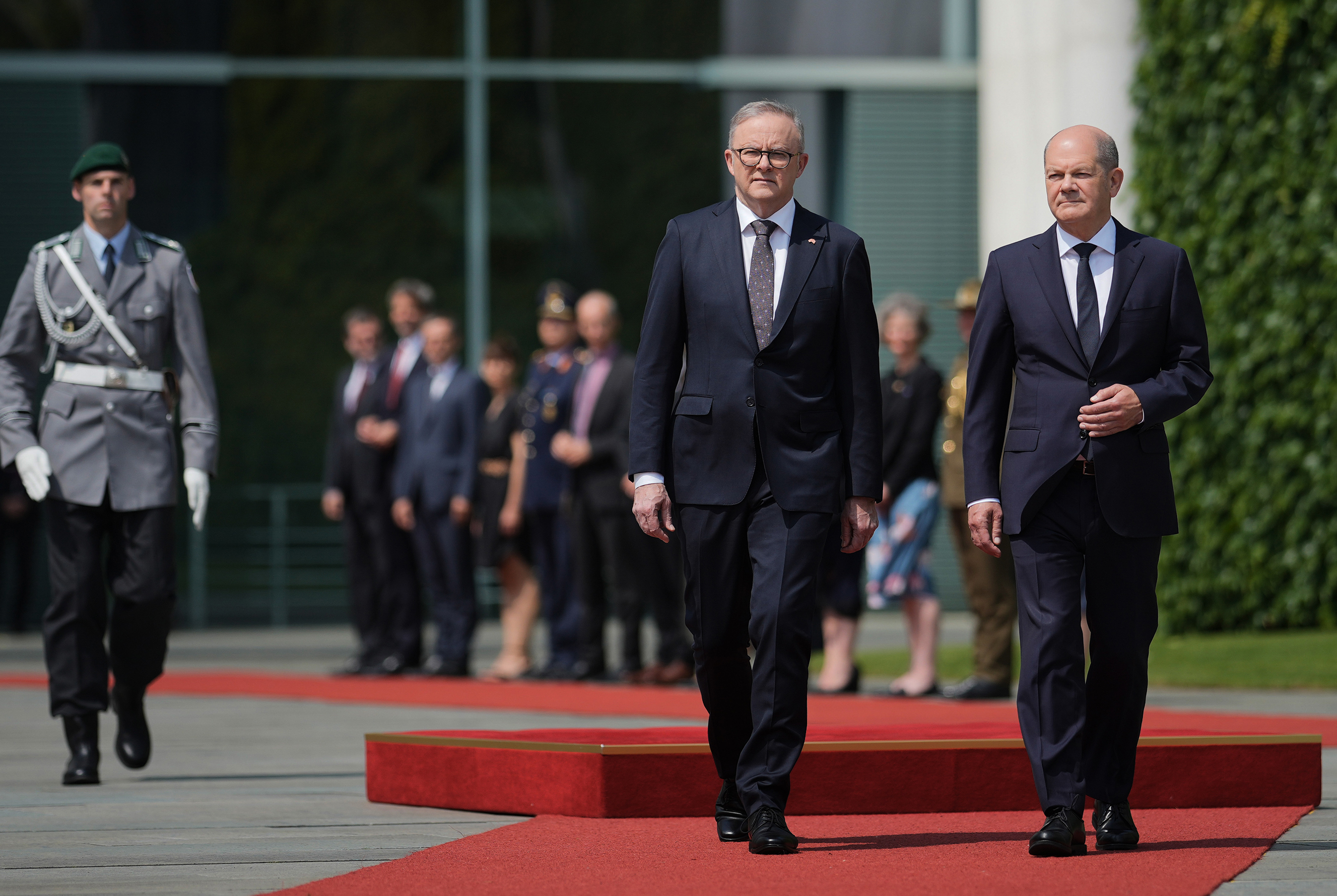 Two men in suits walk down a red carpet with decorated soldiers. 