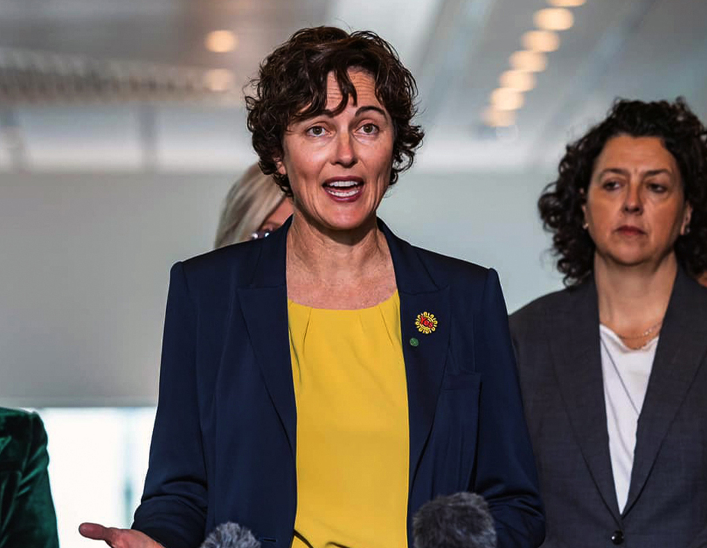 A woman in a yellow top and navy blue blazer speaks at a press conference.