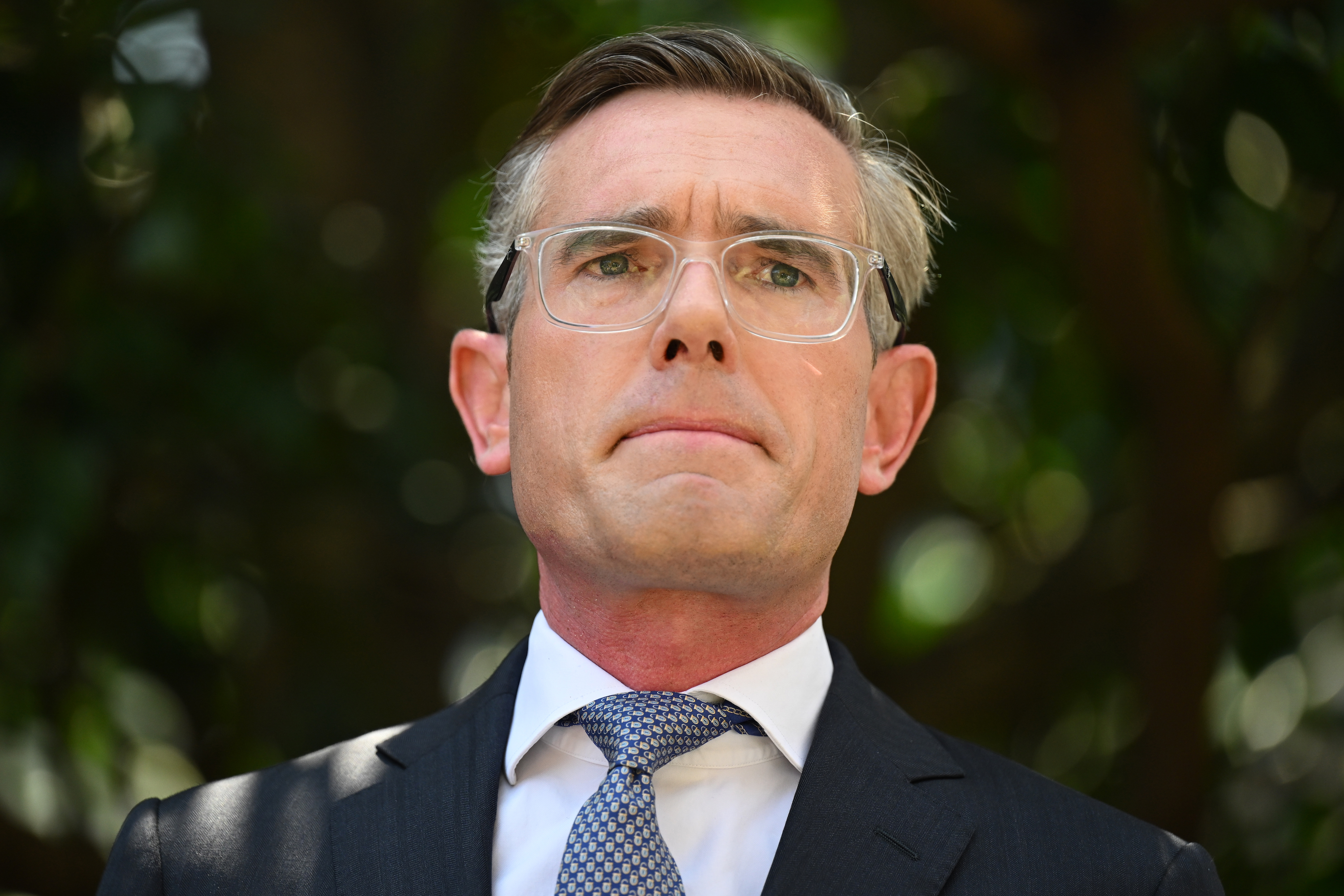 NSW Premier Dominic Perrottet; a 40-year-old Caucasian man with short greying hair wearing clear-rimmed rectangular glasses. The image is a closeup of him looking extremely contrite with his lips pressed together.