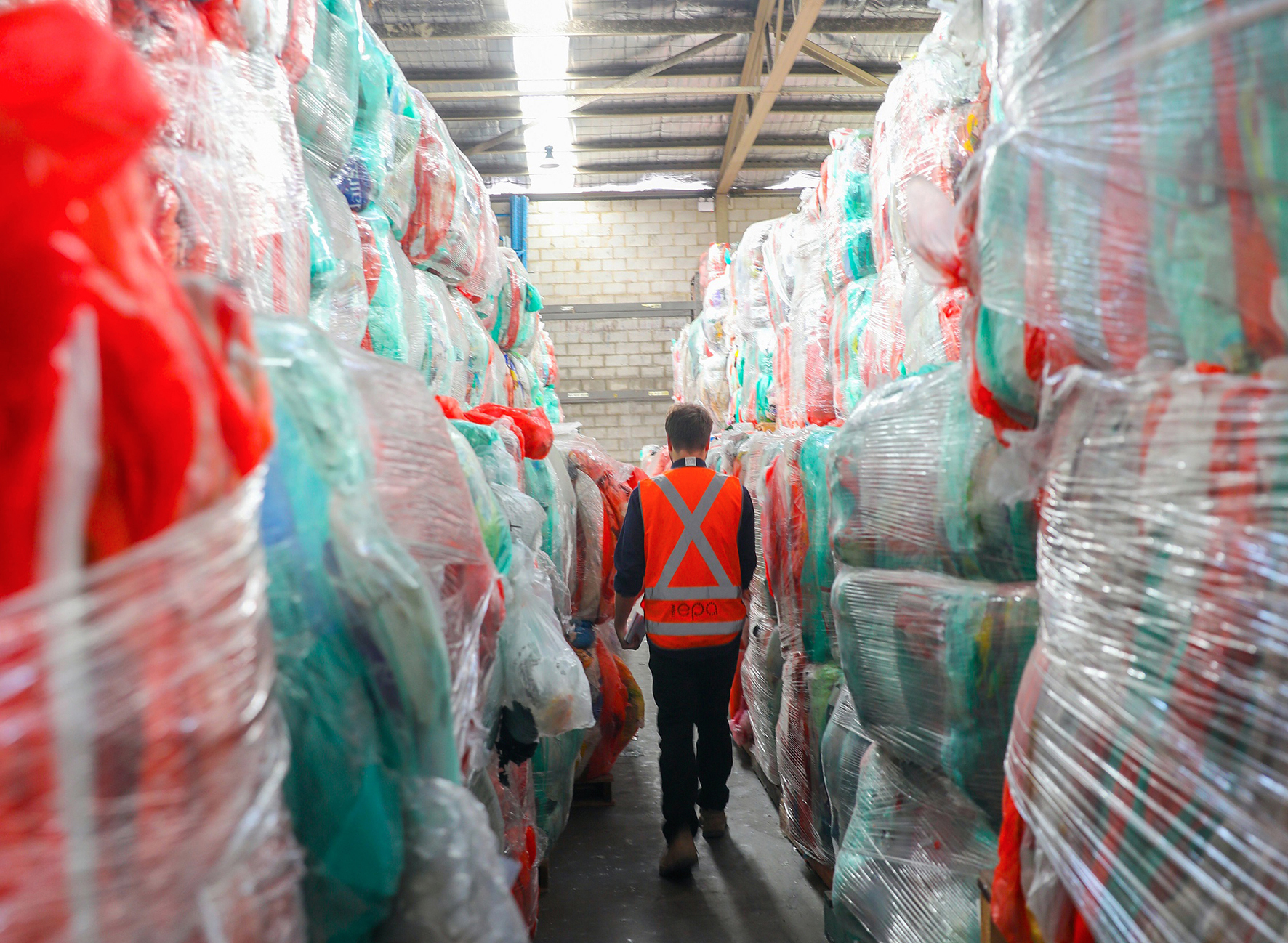 A man in a high-vis vest walking between bags of soft plastic in a large warehouse.