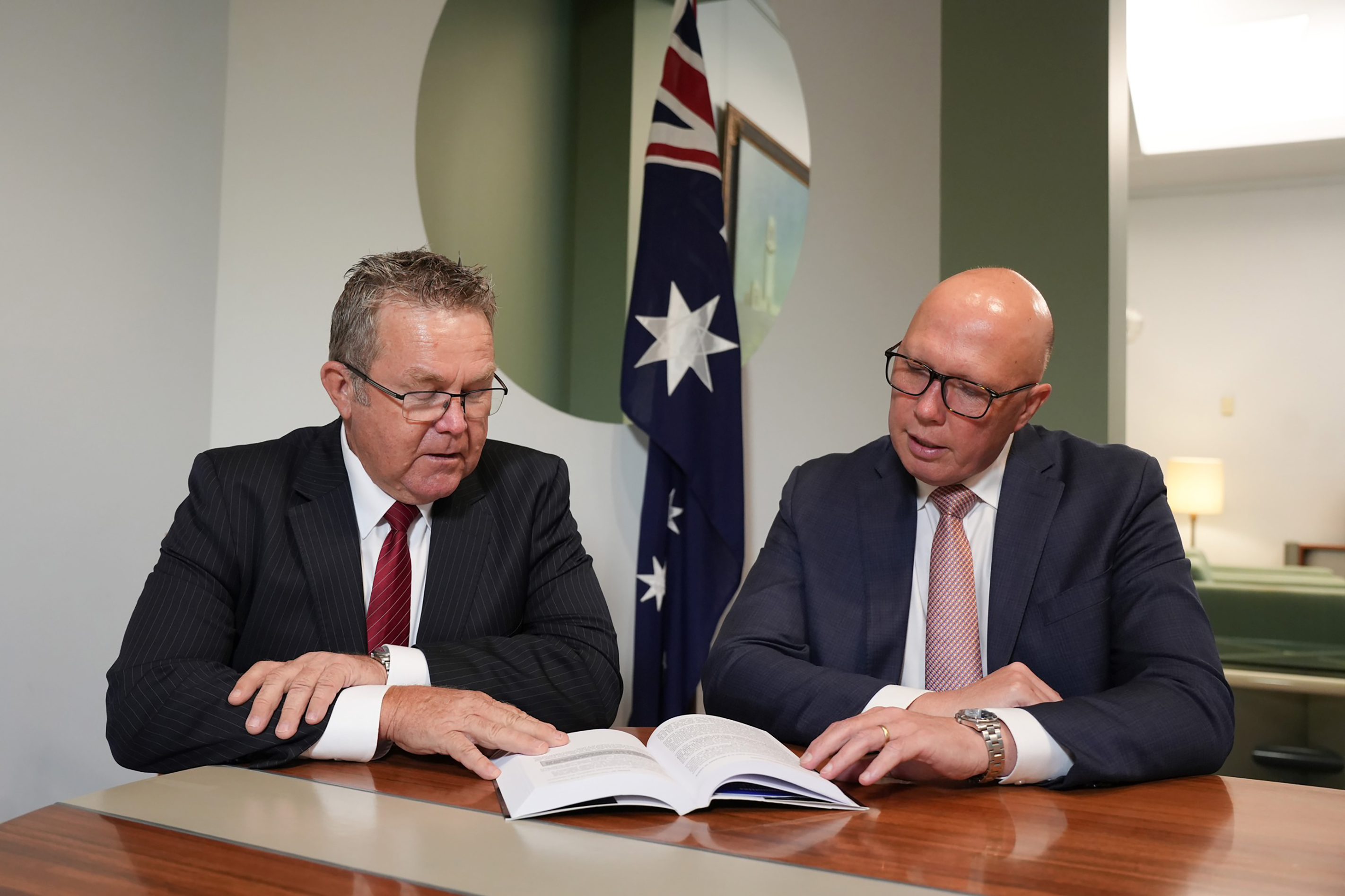 Peter Dutton and Colin Boyce sit at a desk and read from the page of an open book. An Australian flag stands behind them.