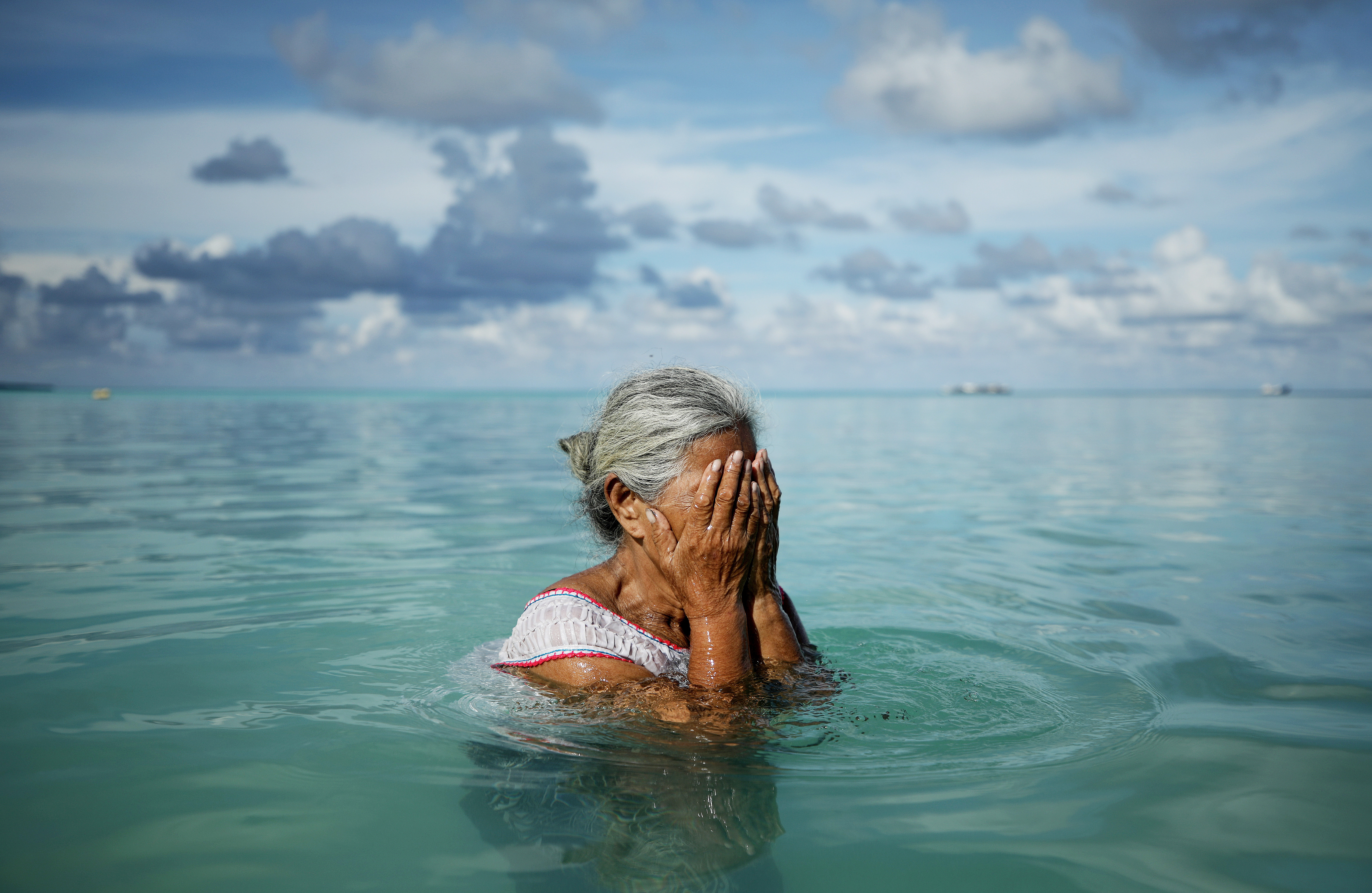 A woman with grey hair stands in crystal blue water with her hands covering her face.