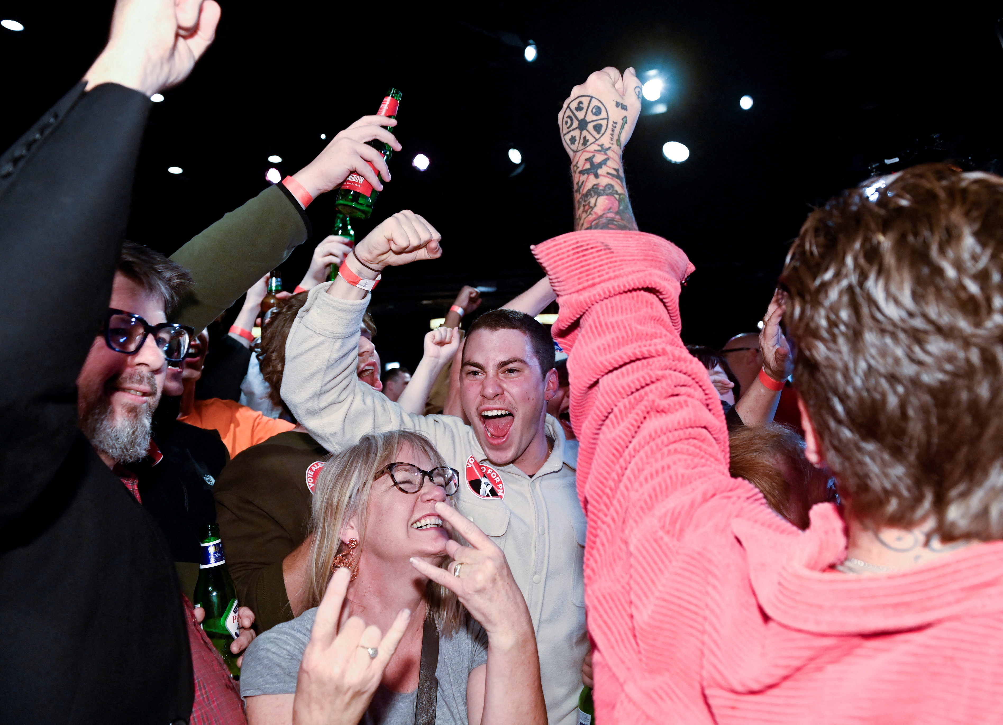 A group of people in a crowd are illuminated by a flash, they are obviously celebrating and yelling with beers in hand and jubilant expressions.