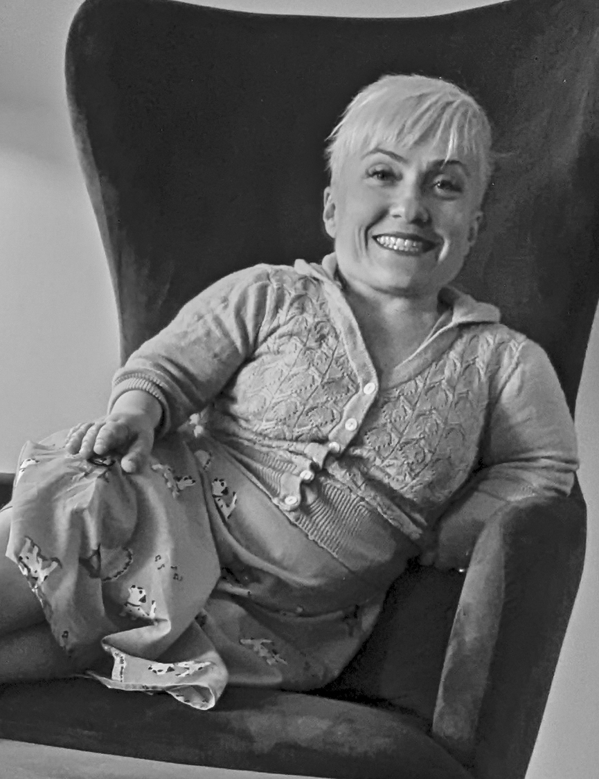 A black and white image of a Caucasian woman with dwarfism sitting on a winged armchair smiling at the camera. She is wearing a button up shirt and her hair is cropped short in a pixie cut. 