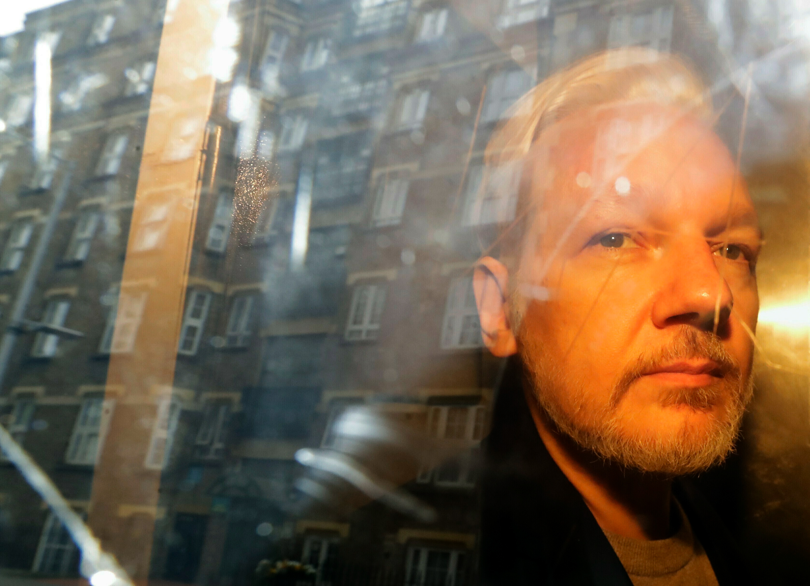Julian Assange gazing through a car window. The window catches the reflection of London apartments.