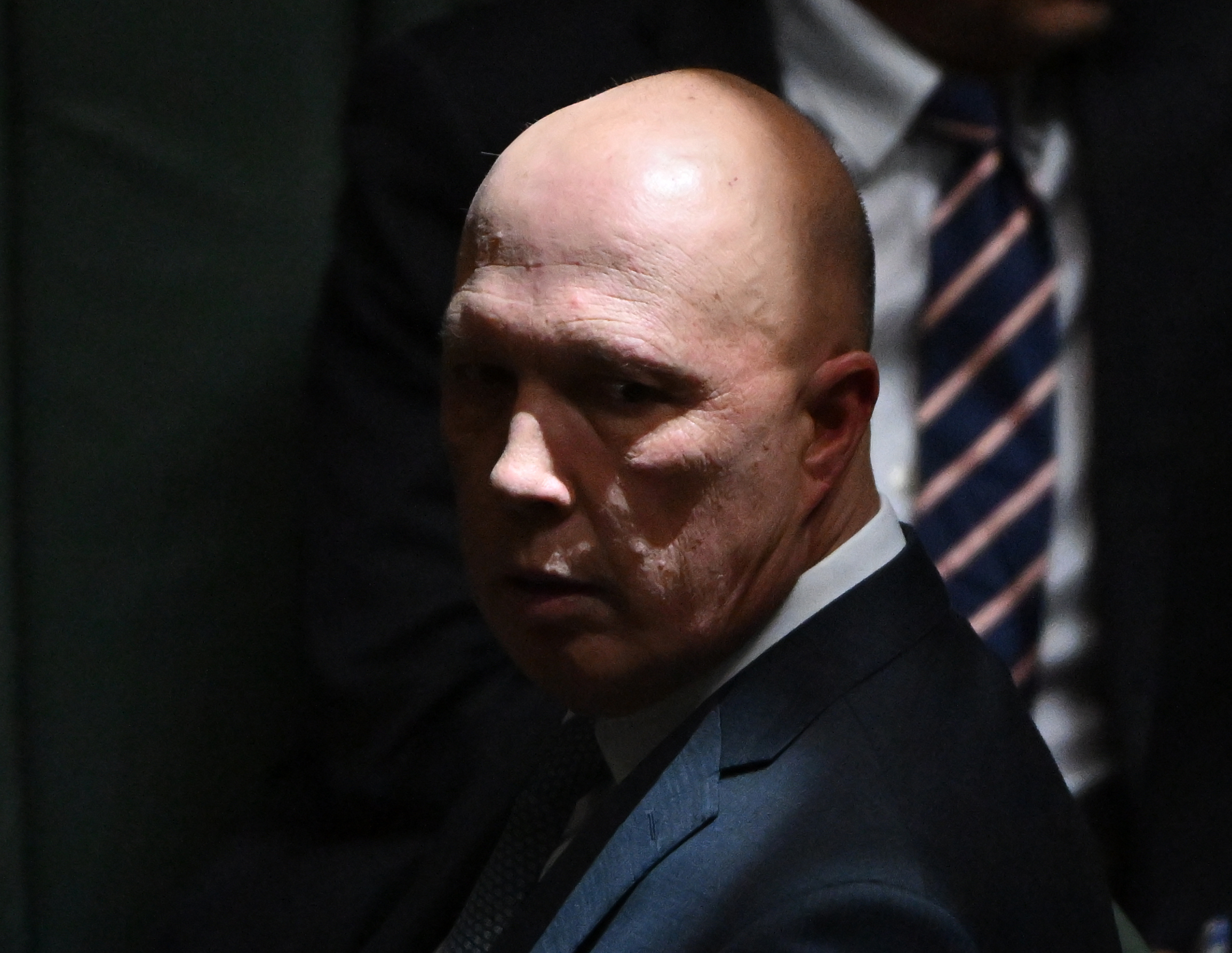 Opposition Leader Peter Dutton in parliament - he is a Caucasian man with a bald head and stern expression wearing a dark blue suit. He is looking to his left and there is a ray of light on his head, with the rest of the image in shadows.