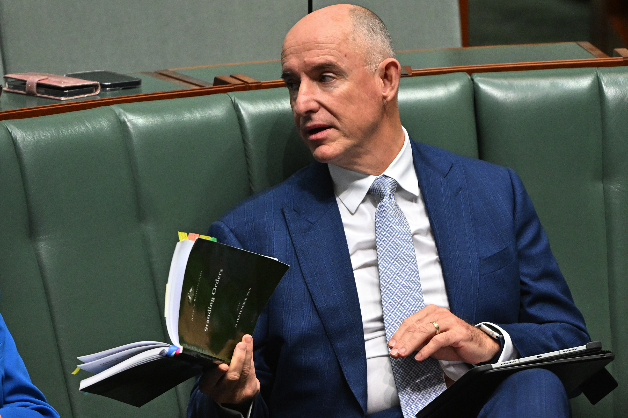 A middle-aged man sits in parliament with an open book raised in his hand.