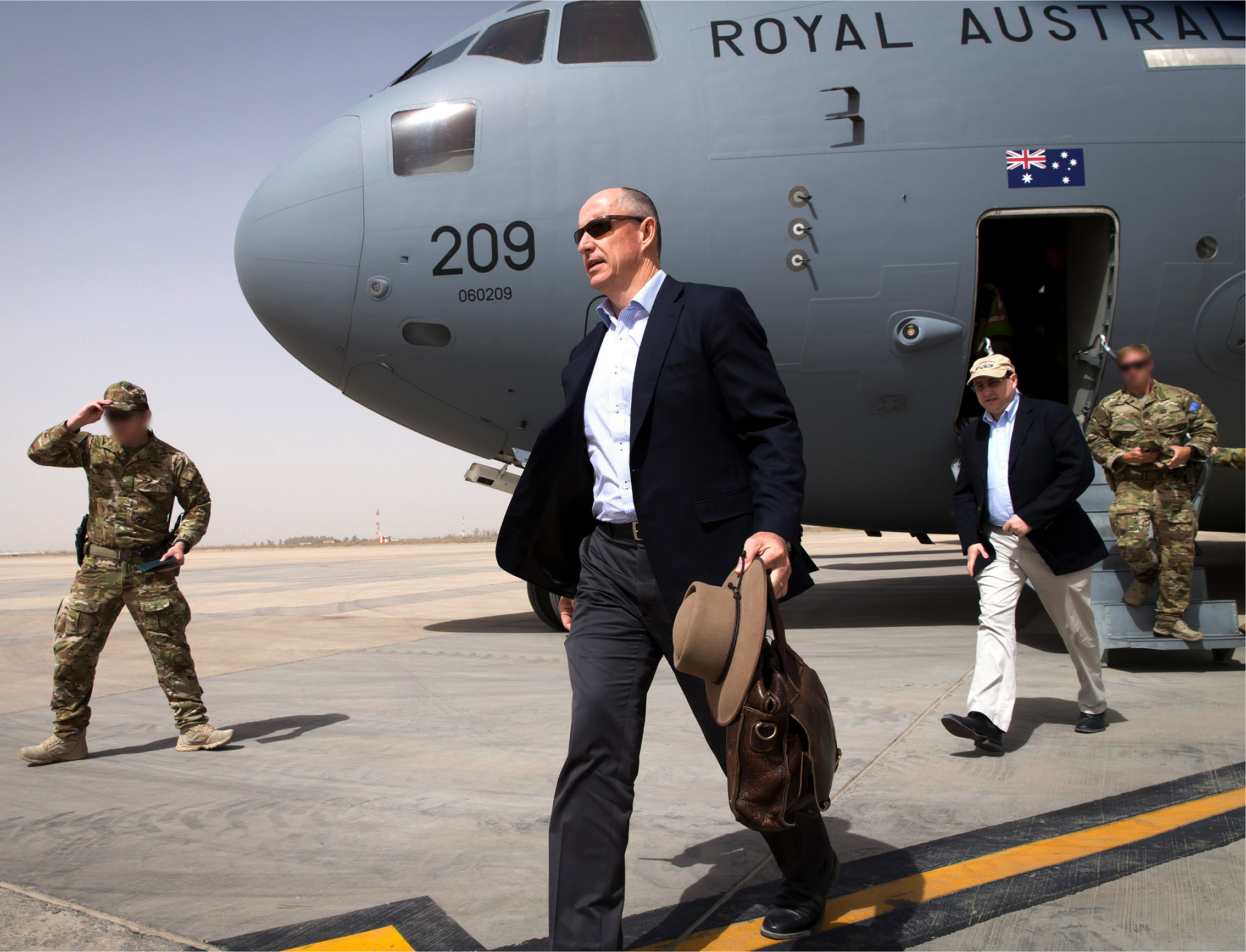 A man carrying a leather bag walks across an airplane runway with men in military uniform and a plane behind him.