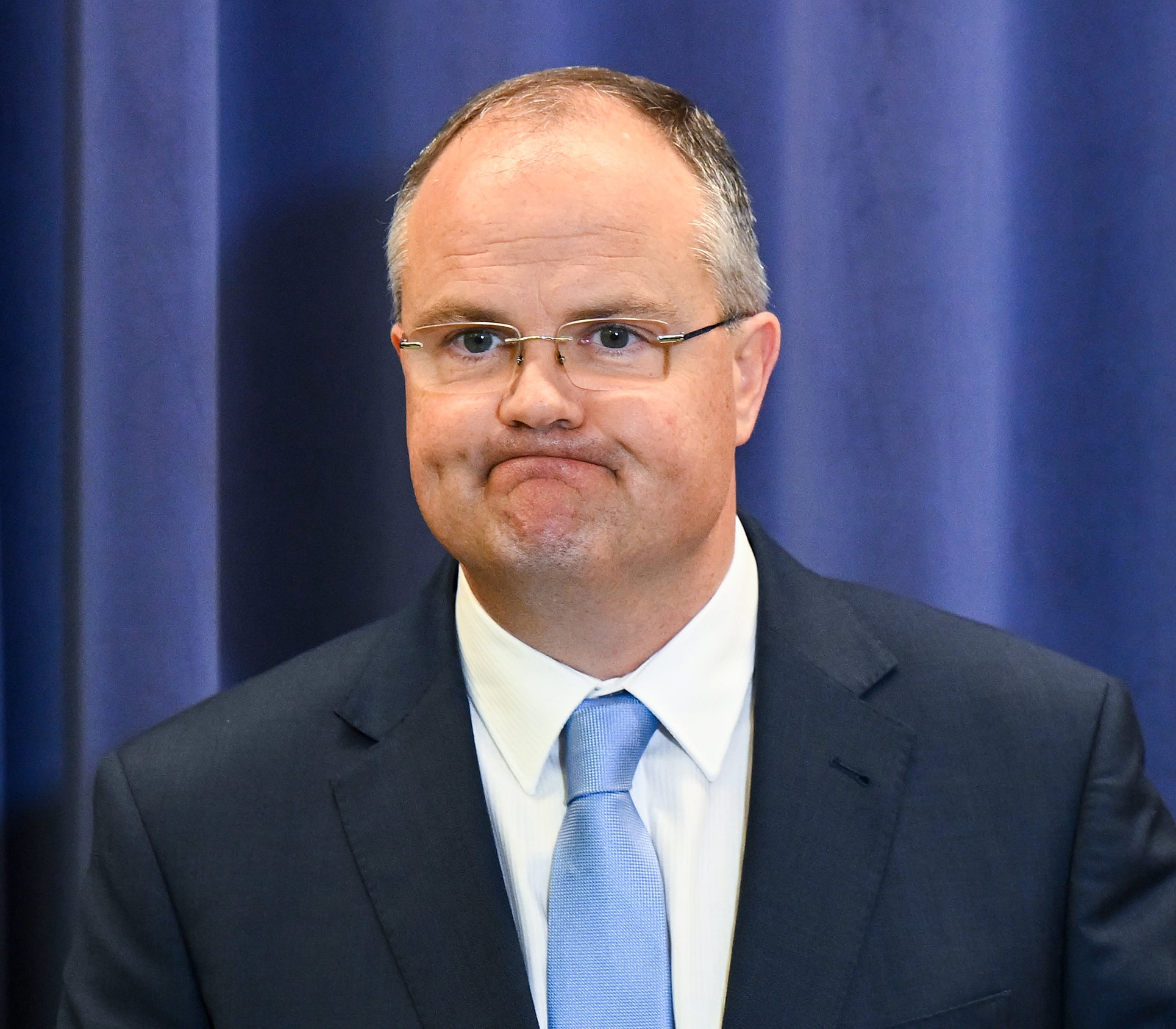 A blading man in a suit stands in front of a blue curtain.