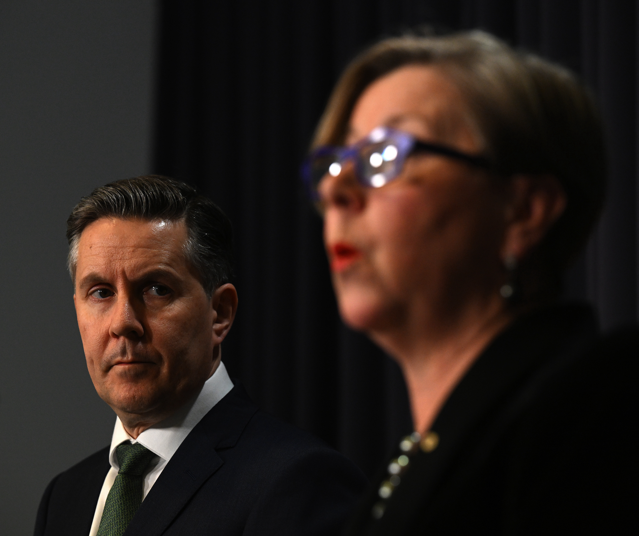A Caucasian man with brown hair going grey in his forties, wearing a formal suit and green tie is in focus looking on to a woman out of focus to the right of the frame, who appears to be speaking to the media in front of a microphone