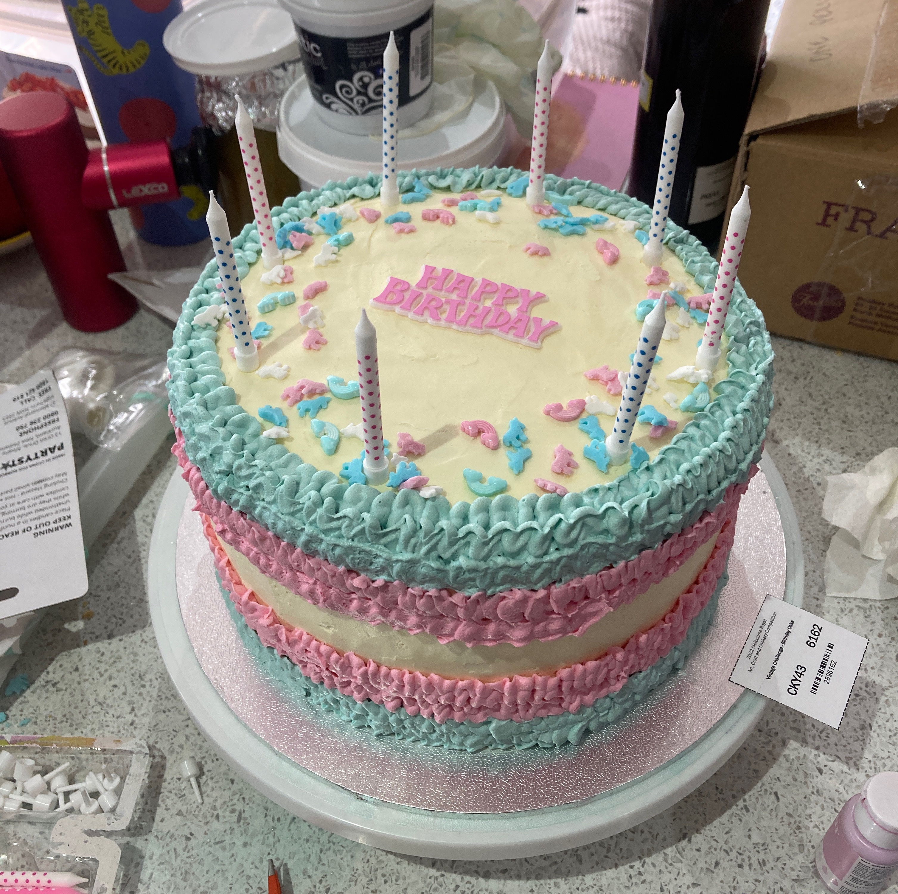 Amid a kitchen bench mess of cooking paraphernalia, a teal, pink and cream birthday cake with "Happy Birthday" and eight candles sits in the centre. It has multi-coloured icing around the sides of the cake and is beautifully garish. 