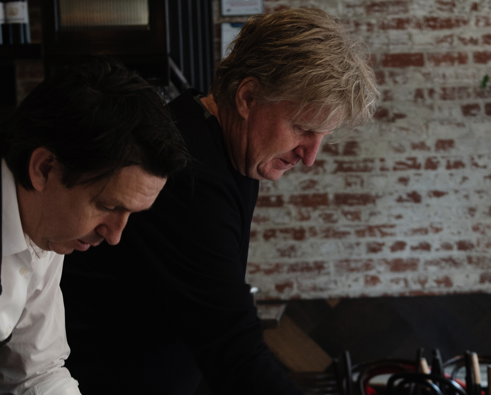 Two men lean over a kitchen table.
