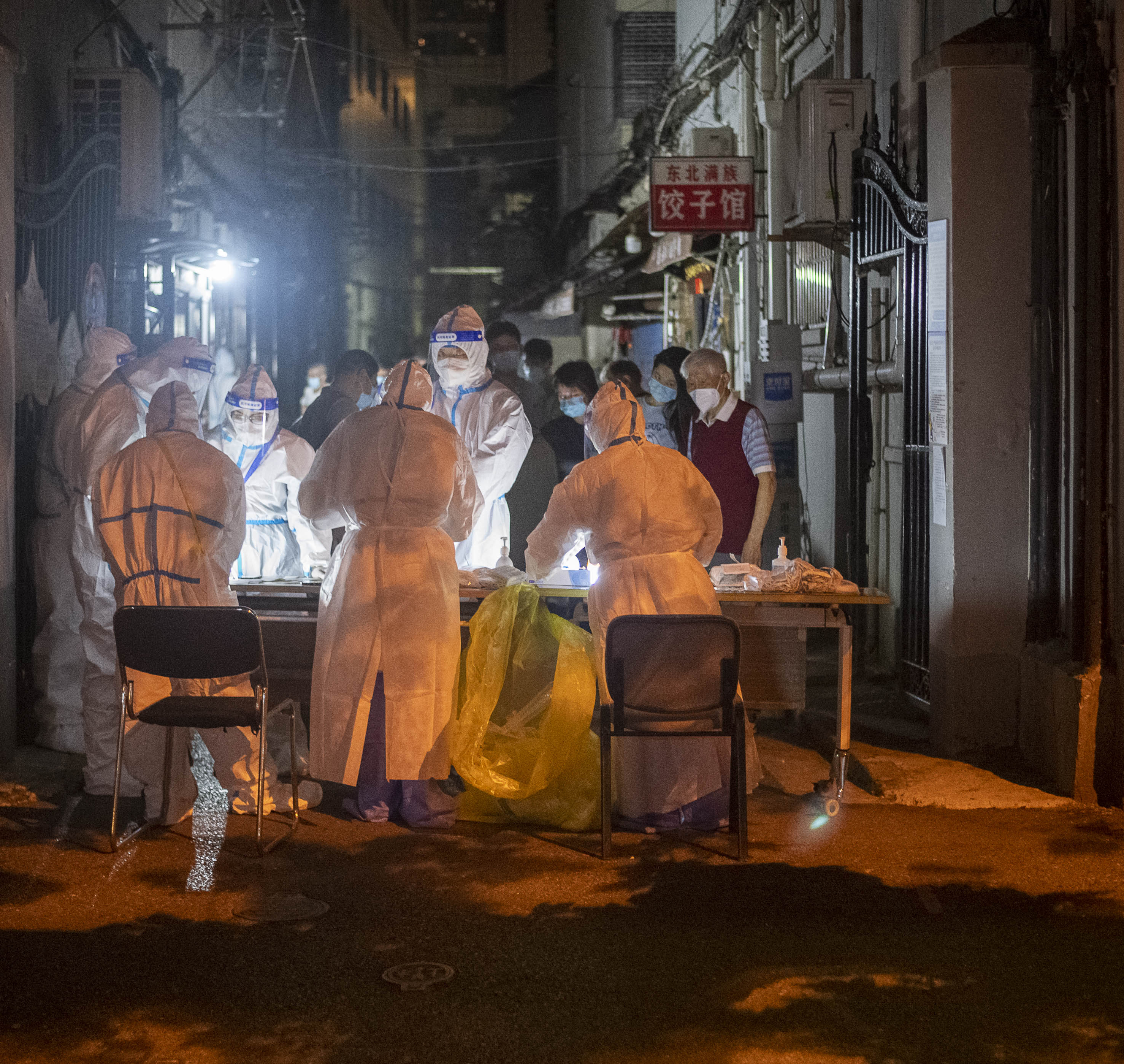 Several people dressed in hazmat suits surround a table on a Shanghai street, organising Covid-19 tests. Several onlookers wearing masks watch in the background. 