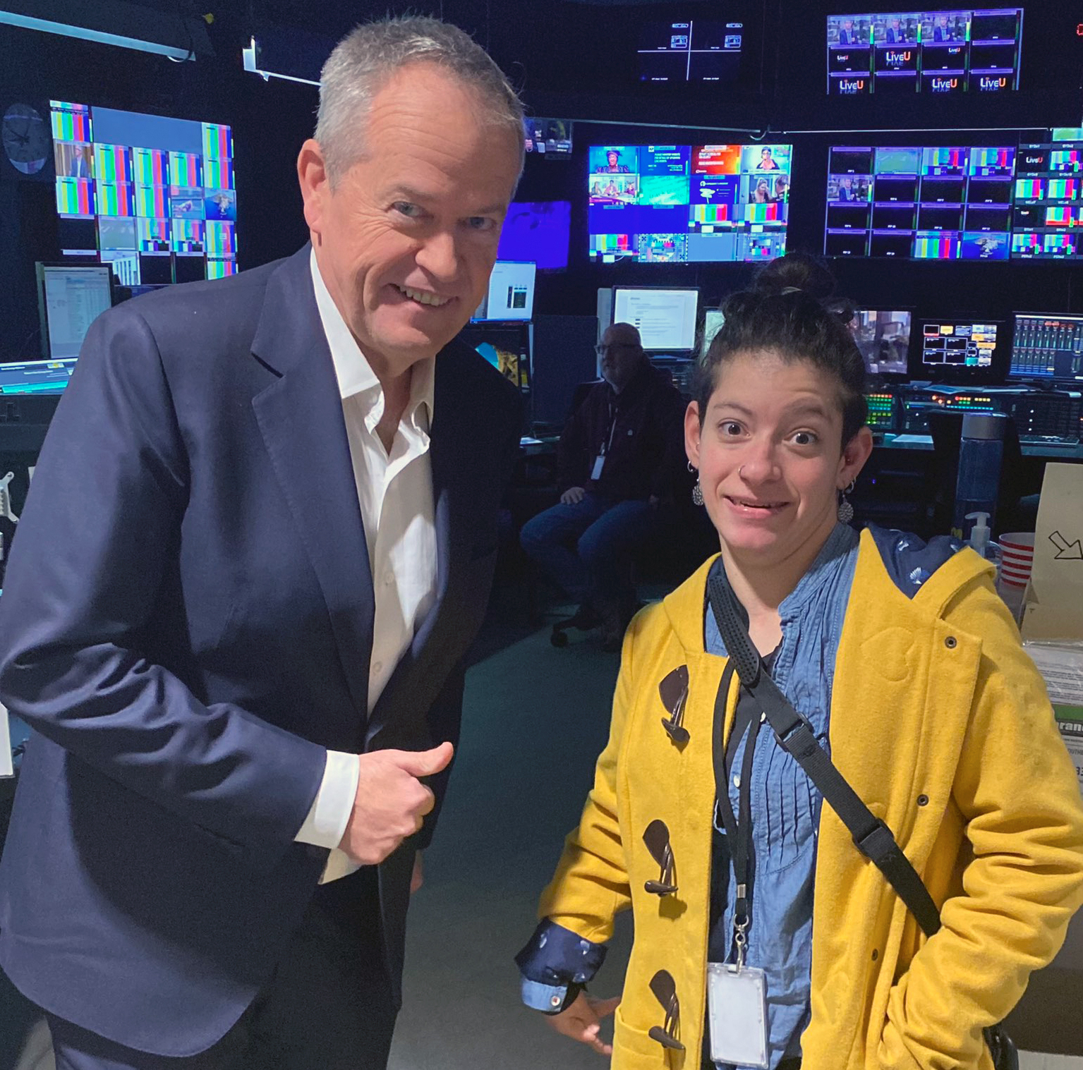 Two people are in a television production studio, with lots of blue screens behind them. The man on the right is middle-aged and Caucasian, wearing a blue suit and white shirt with a smile and his hand doing a "thumbs-up". The person on the right is a younger woman wearing a bright yellow pea coat, a blue denim shirt, silver earrings and a nose-ring, and her hair pulled back in a bun. She is also smiling. 