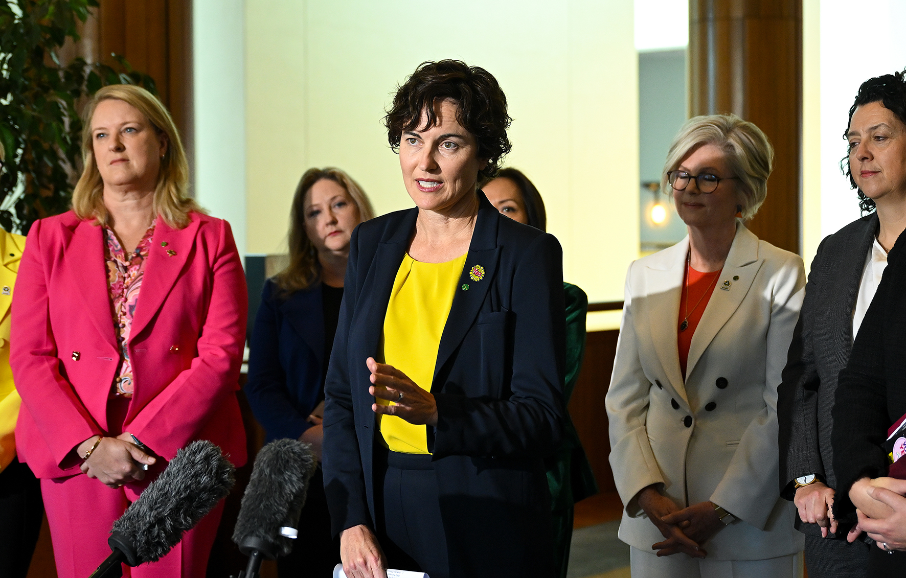 A group of women dressed in blazers stand together at a press conference.