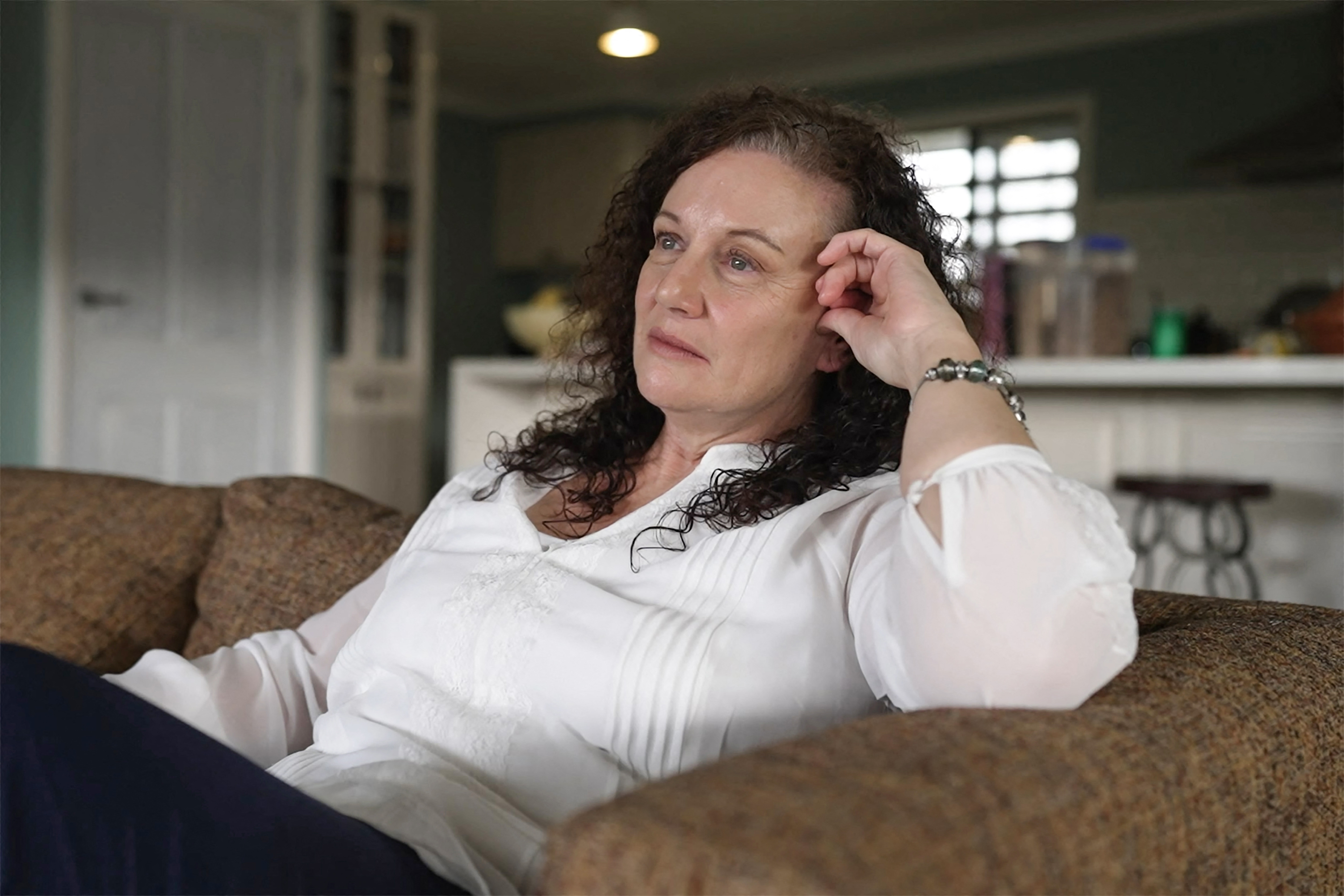 A middle-aged woman with dark hair sits on a couch, her hand by her face as she gazes at something out of shot.