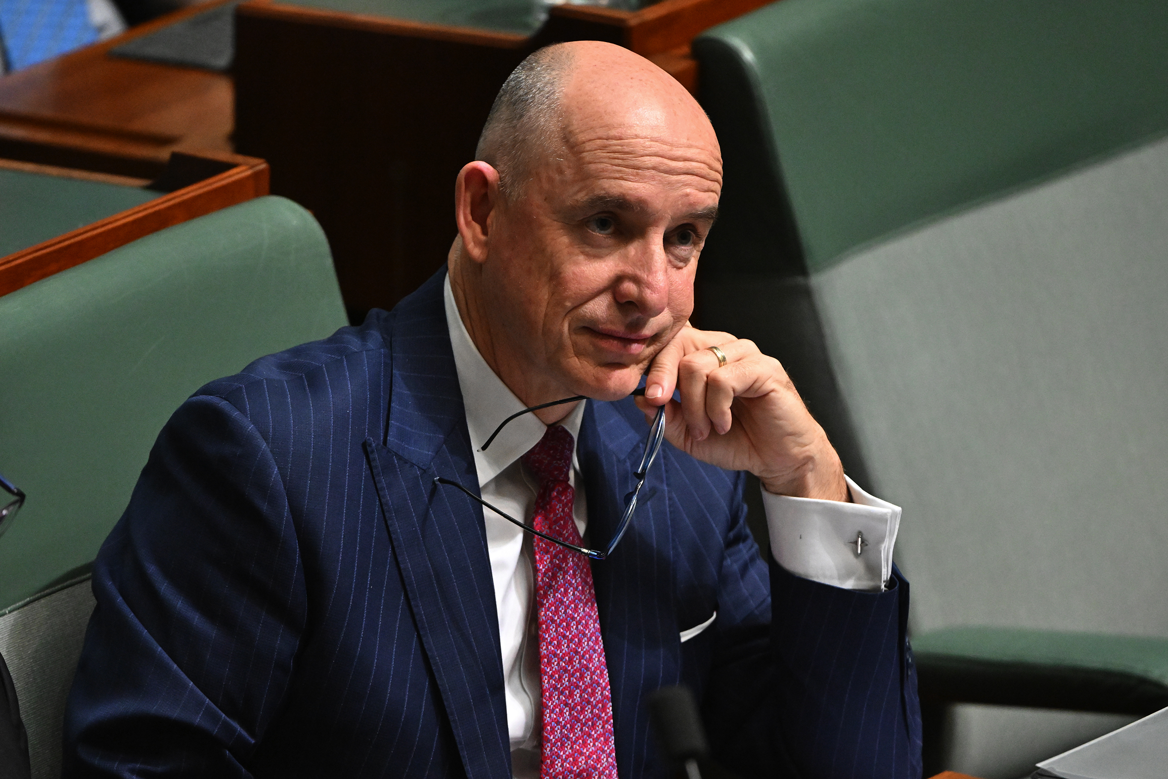 A bald, middle-aged man dressed in a suit sits in Parliament with his glasses in hand.