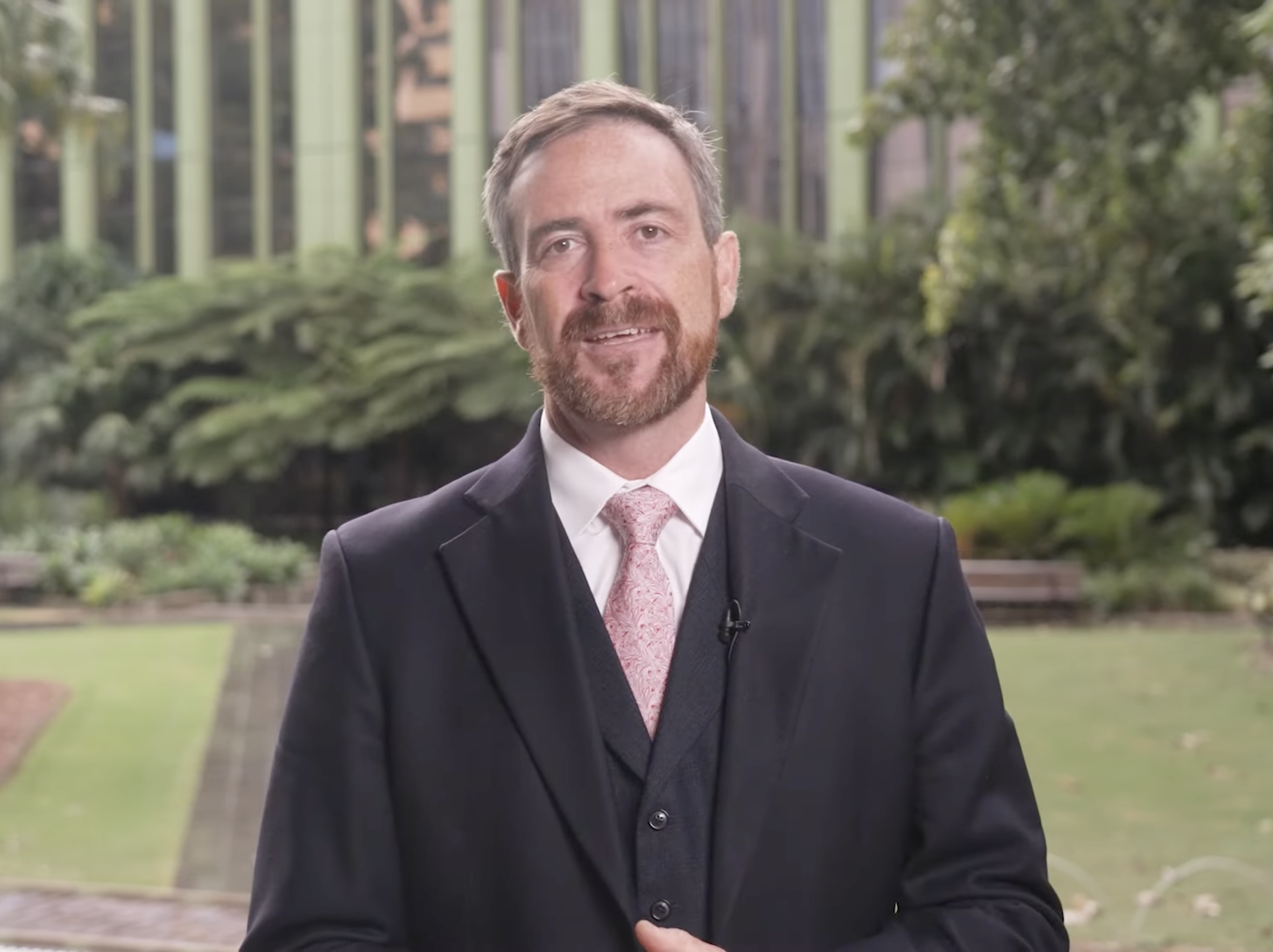 A man wearing a black suit with a garden behind him.