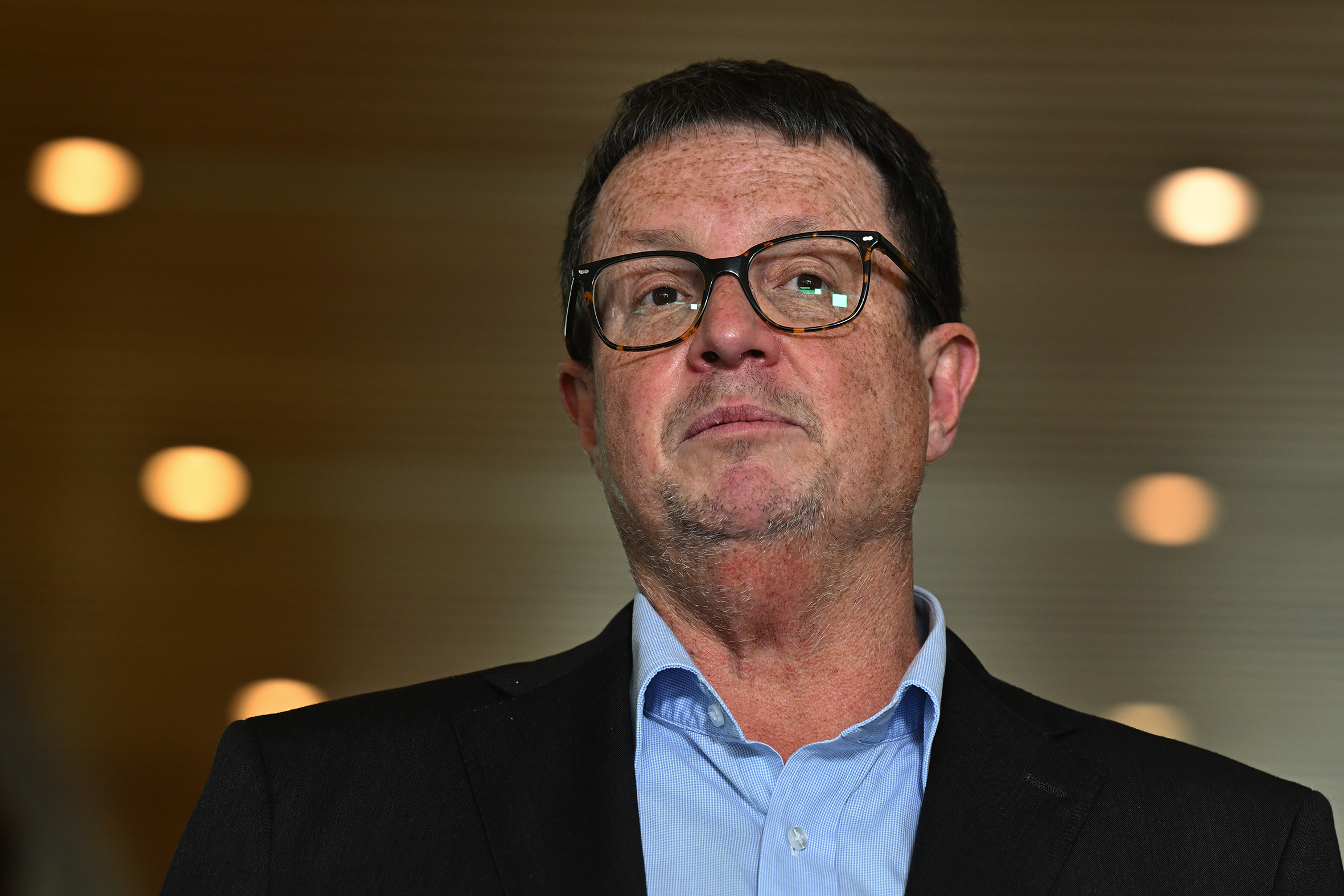 A closeup of a middle-aged Caucasian man wearing tortoiseshell glasses, with brown hair flecked with grey and a lined and unshaven face. He is wearing a blue shirt and dark grey suit jacket.