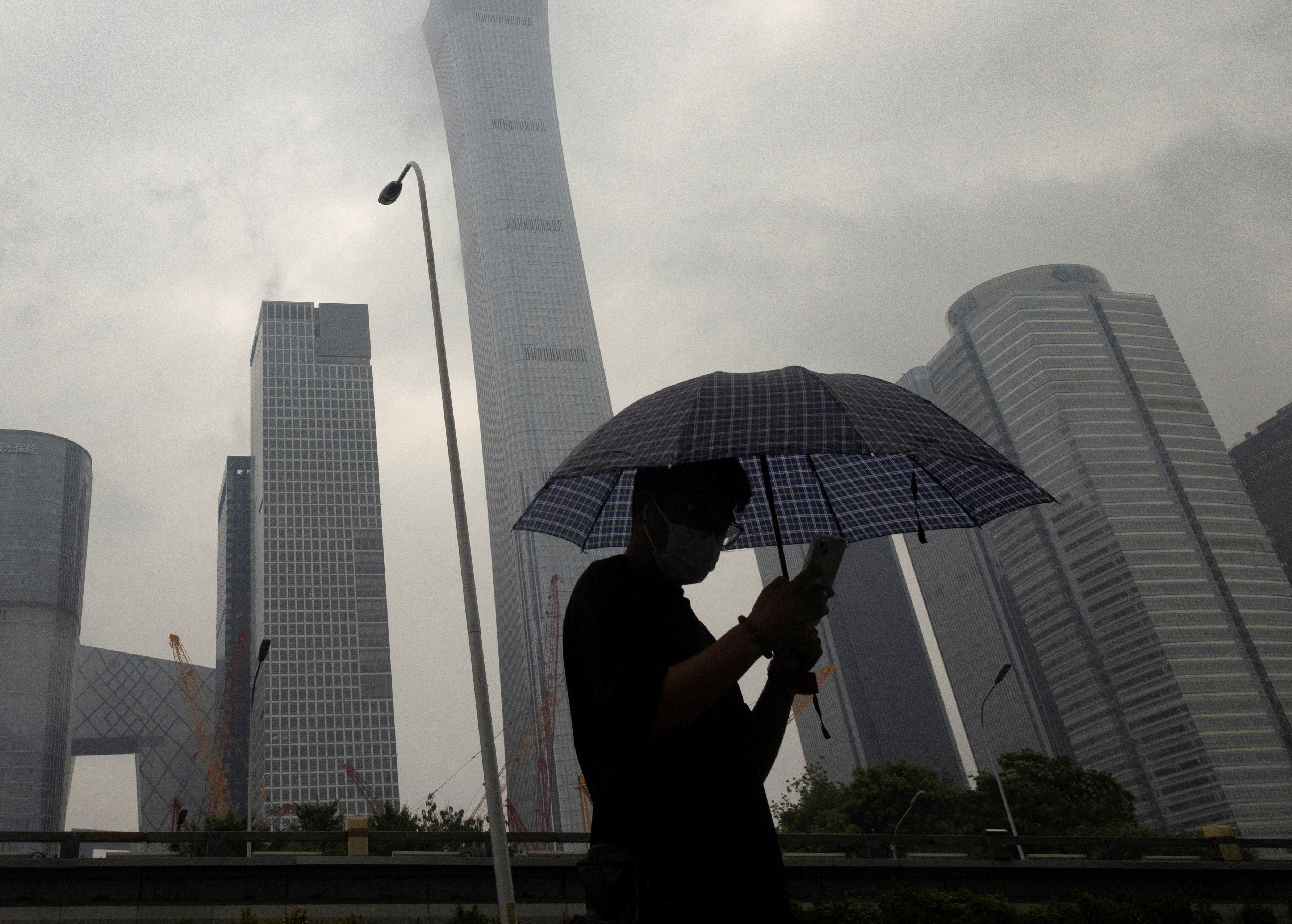 Skyscrapers under an overcast sky. In the foreground, a man wearing a mask and holding an umbrella.