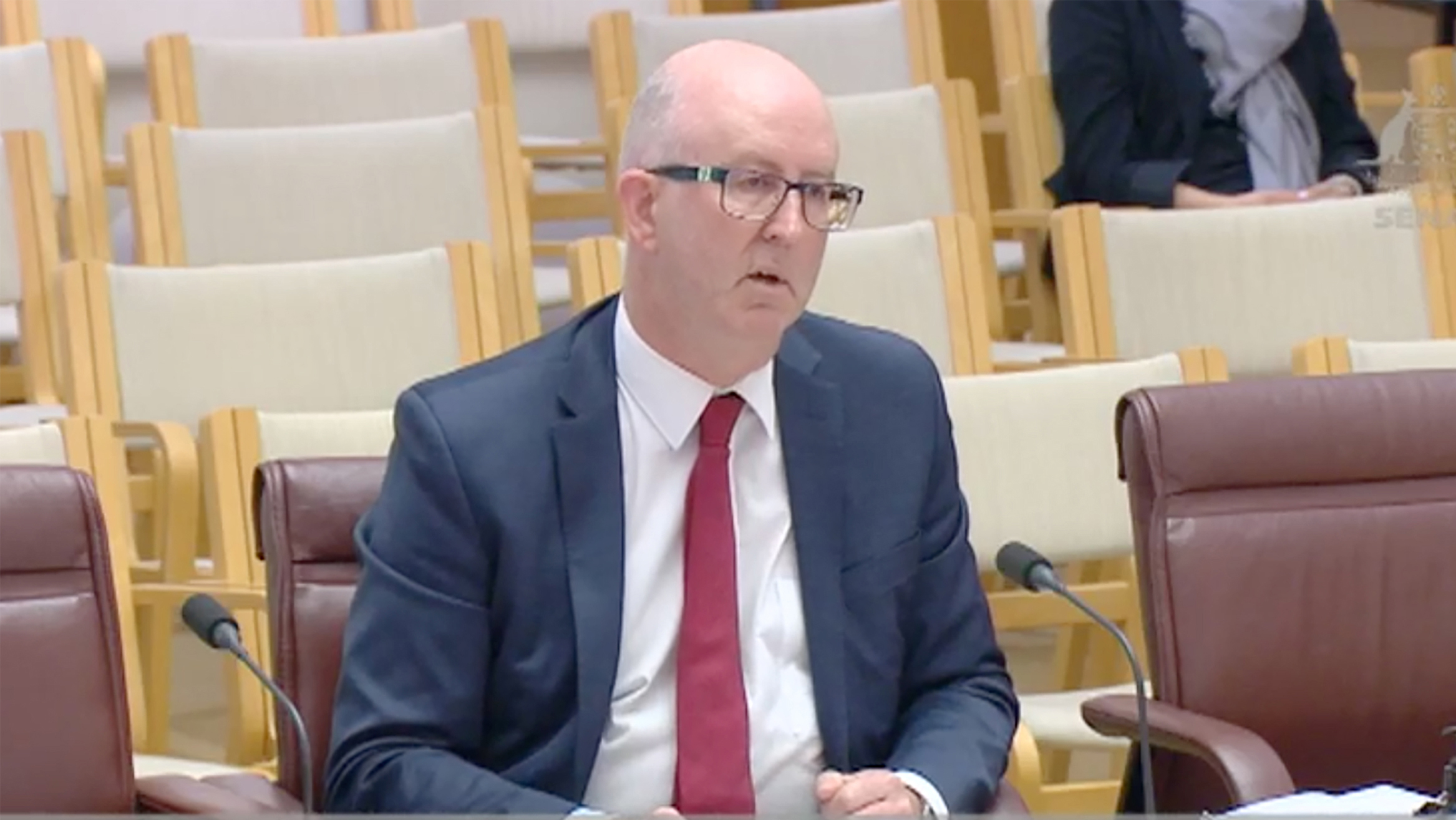 A bald man wearing glases and a suit sits by a microphone.