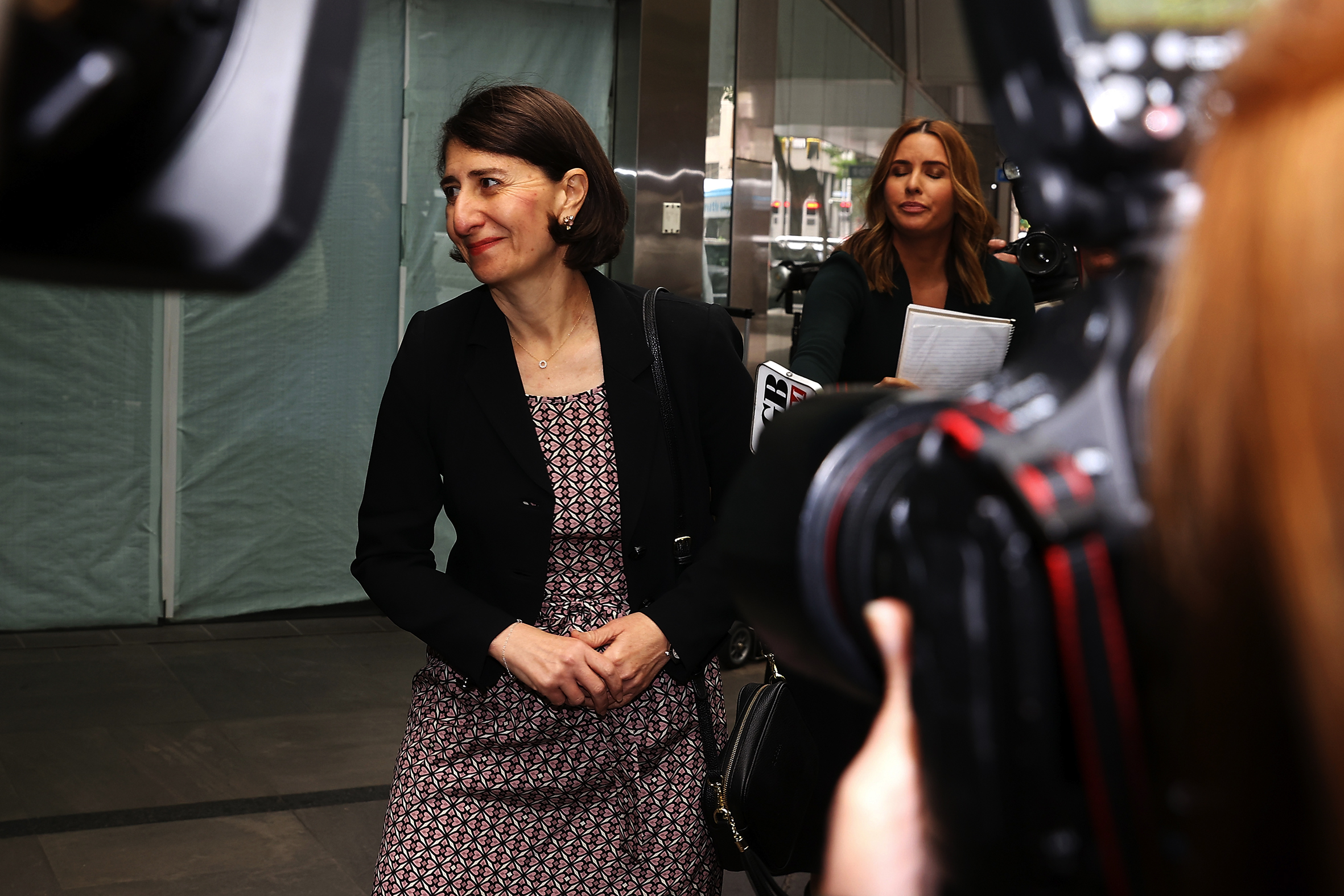 A middle-aged woman on a street footpath and tailed by journalists holding cameras and microphones.