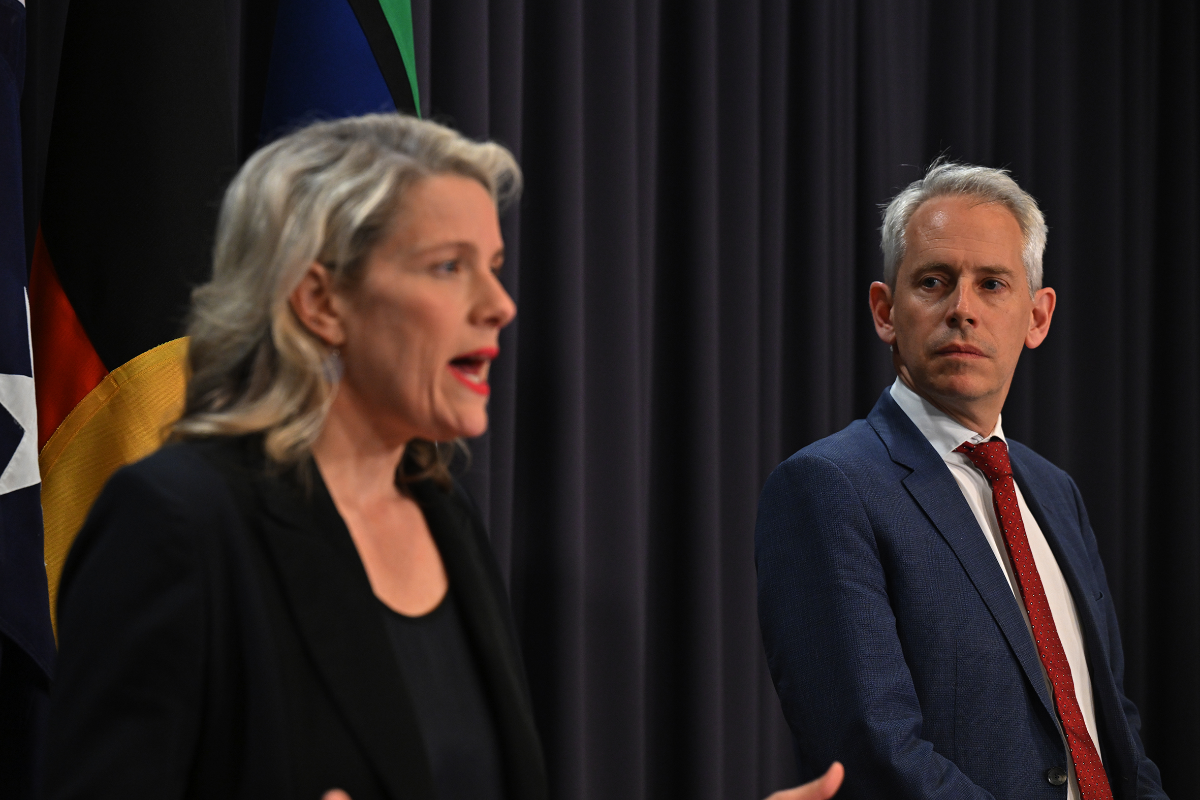 A woman in a black blazer speaks while a man with grey hair and dressed in a navy suit stands watching beside her. 